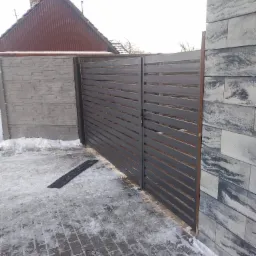 Nowoczesna brama wjazdowa z poziomych, szarych profili metalowych, zamontowana pomiędzy dwoma fragmentami ogrodzenia z betonowych bloczków imitujących kamień, częściowo pokryta śniegiem.