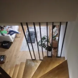 Widok z góry na drewniane schody z metalową balustradą w nowoczesnym domu, w tle salon z wypoczywającą osobą na kanapie.