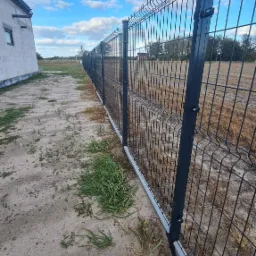 Długi, czarny panelowy płot z betonową podmurówką, biegnący wzdłuż piaszczystej działki z kępami trawy, widok z boku, w tle budynek w stanie surowym i błękitne niebo.