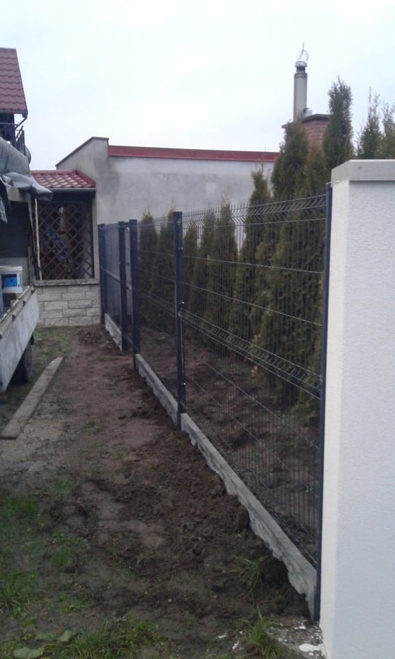 Montaż ogrodzenia panelowego z podmurówką betonową, widoczny fragment posesji z tujami w tle, ziemia wokół ogrodzenia jeszcze nieuporządkowana.