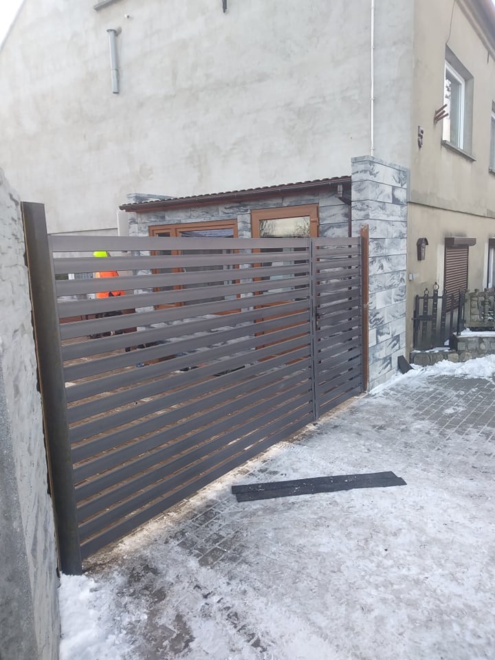 Nowoczesna brama metalowa z poziomymi lamelami w kolorze grafitowym, zamontowana przy posesji z elewacją w odcieniach szarości, na lekko ośnieżonym podjeździe z kostki brukowej.