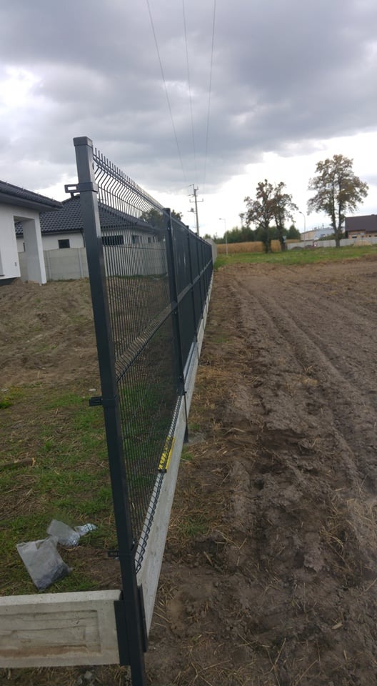 Nowo zainstalowane, ciemnoszare ogrodzenie panelowe z betonową podmurówką na nierównym terenie, widok z boku, w tle budynek mieszkalny i pochmurne niebo.
