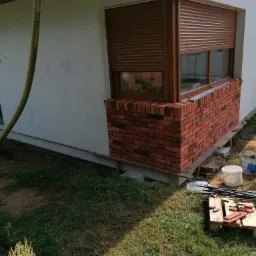 Fragment elewacji domu z oknem z brązowymi roletami i dekoracyjną okładziną z czerwonej cegły klinkierowej w trakcie montażu, widoczne narzędzia i materiały budowlane na trawie.