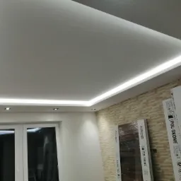 Sufit podwieszany z oświetleniem LED w nowocześnie wykończonym wnętrzu, widoczne okno i fragment ściany wyłożonej dekoracyjną cegłą, na podłodze leżą opakowania po drzwiach.