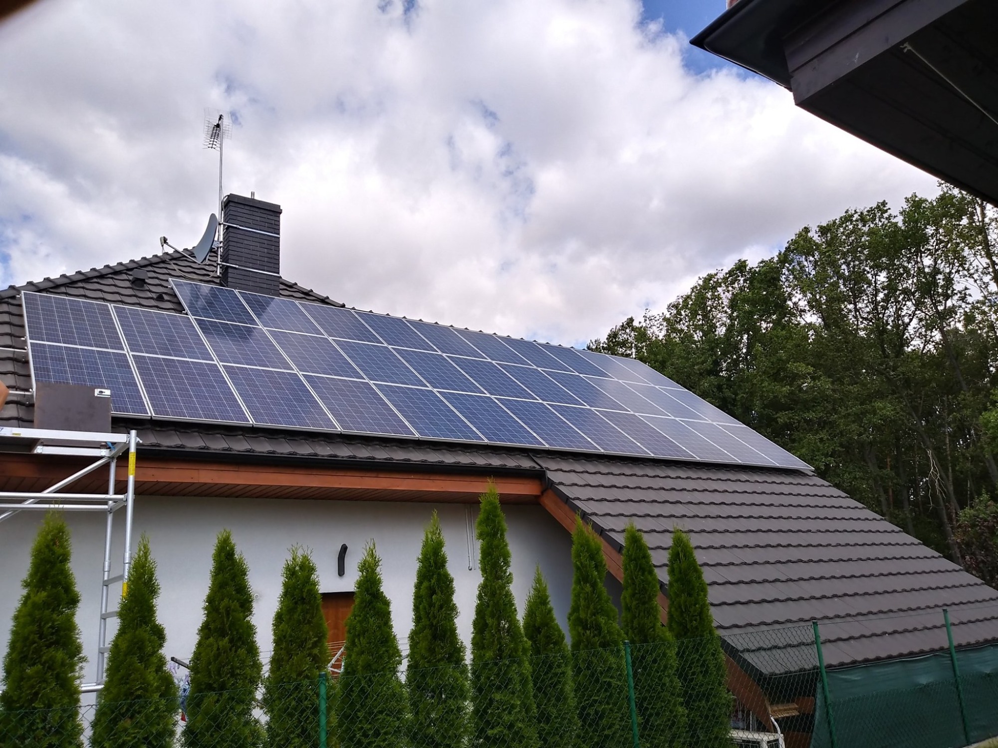Instalacja paneli słonecznych na dachu domu jednorodzinnego z ciemnej dachówki, widoczne rusztowanie i antena satelitarna na kominie.