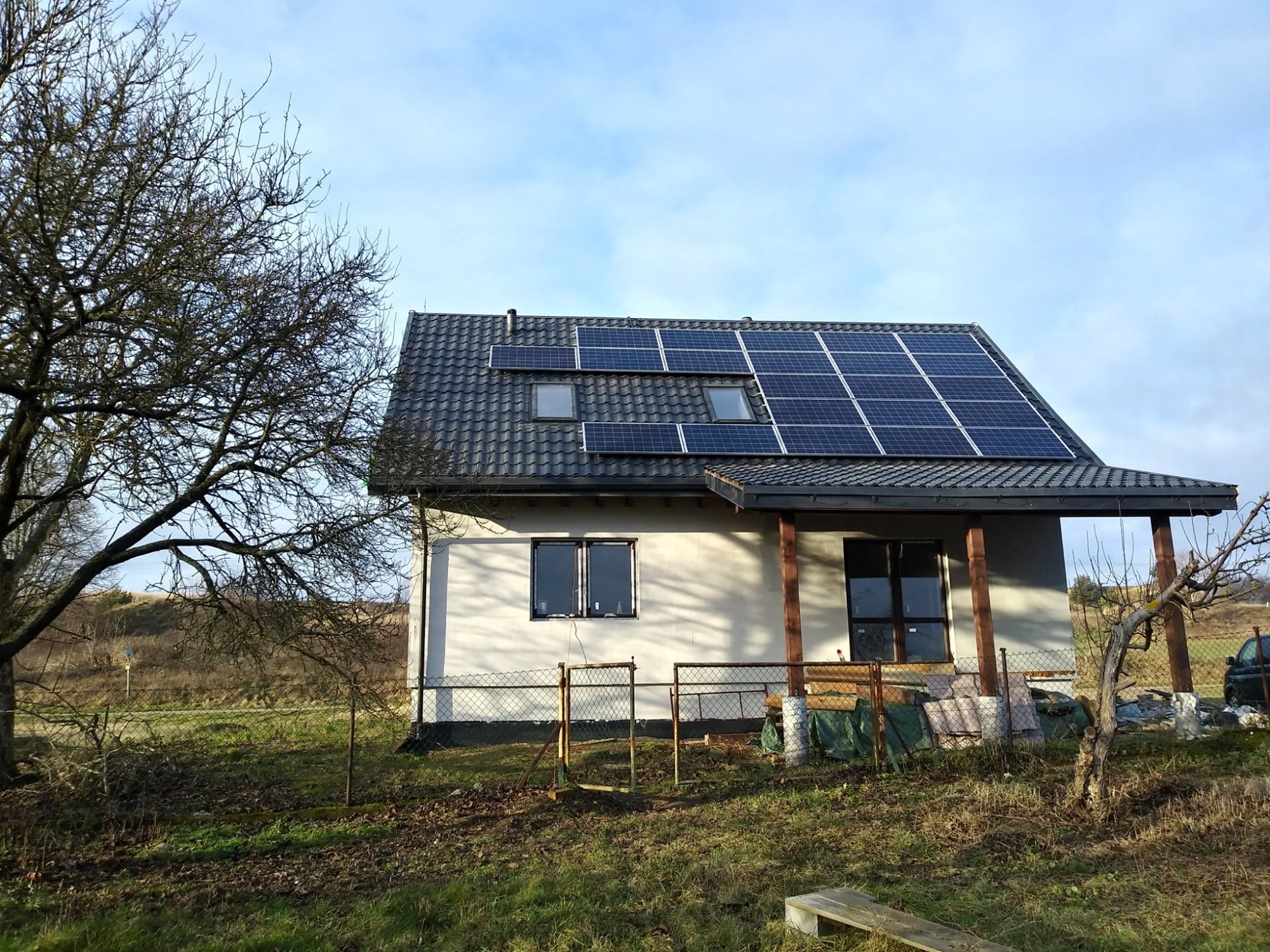 Dom jednorodzinny z panelami słonecznymi na dachu, widoczne okna dachowe, drzewo bez liści z lewej strony, metalowe ogrodzenie i trawa na pierwszym planie.