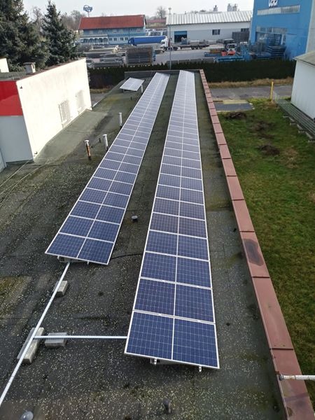 Dwa rzędy paneli słonecznych na płaskim dachu przemysłowego budynku, widoczne elementy montażowe i tło z zabudowaniami fabrycznymi.