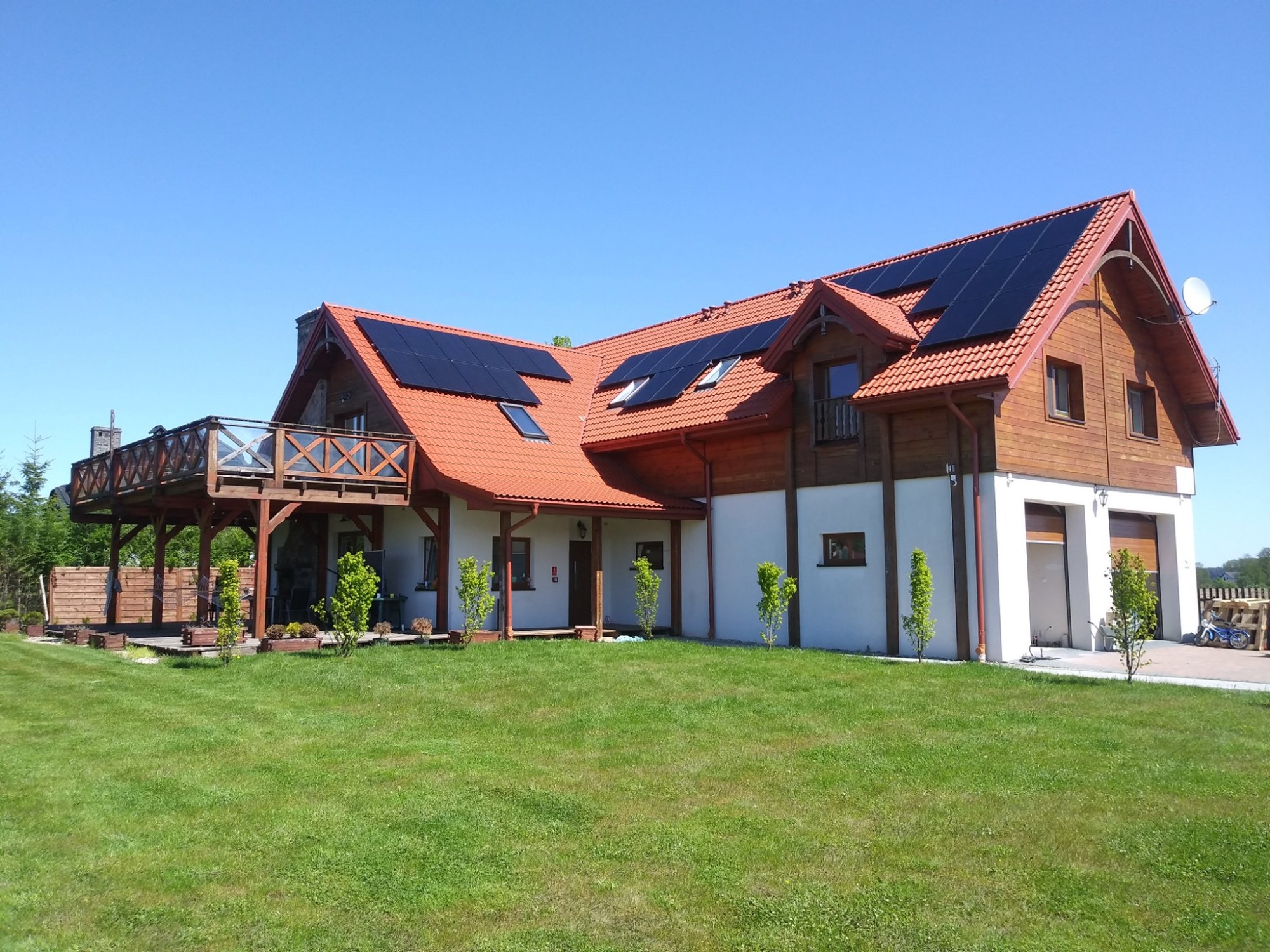 Duży dom jednorodzinny z czerwonym dachem i panelami słonecznymi na kilku połaciach dachu, widoczne z poziomu zadbanego trawnika w słoneczny dzień.