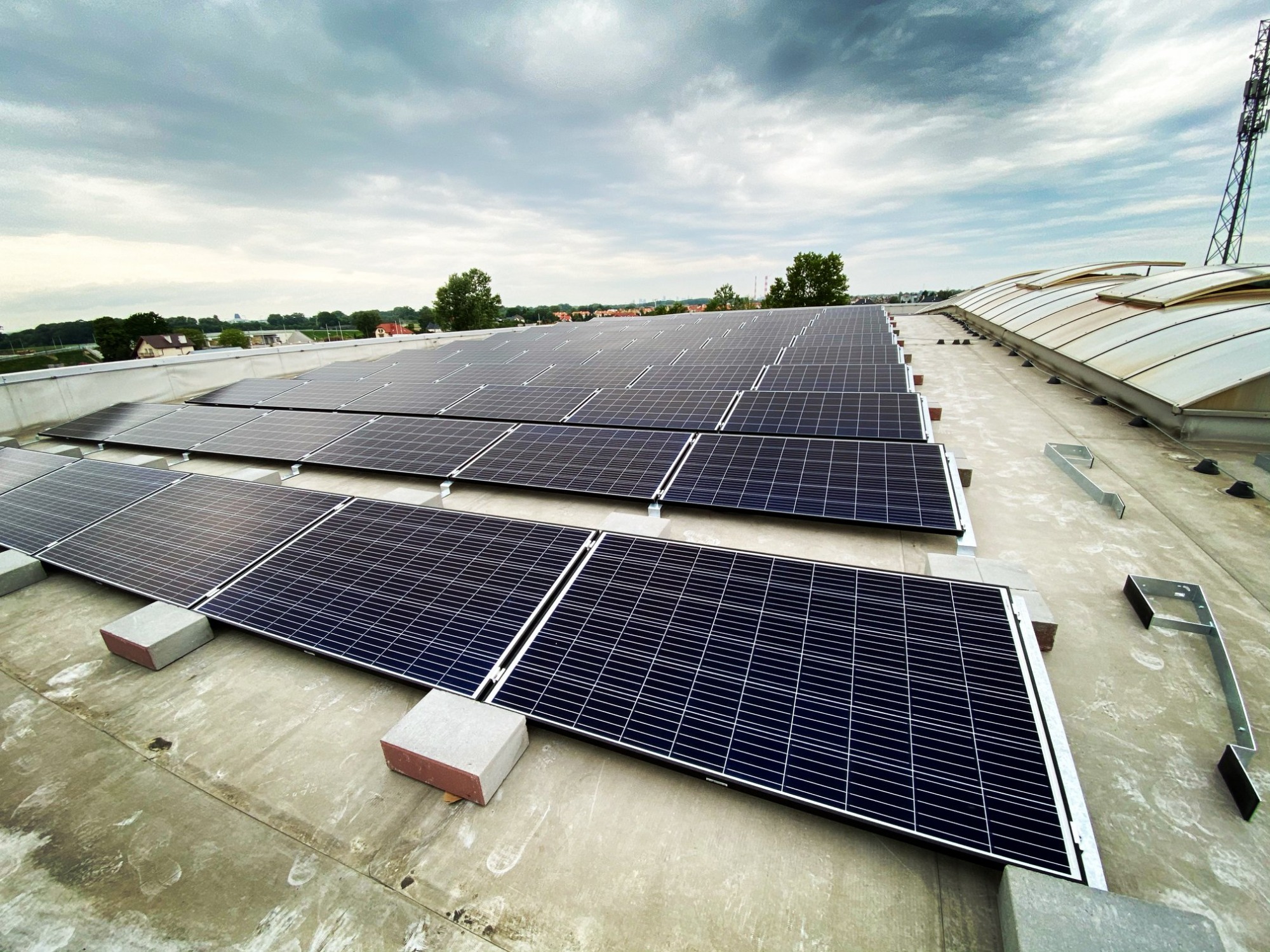 Rząd paneli słonecznych na płaskim dachu, zamontowanych na betonowych blokach, z widocznymi elementami konstrukcyjnymi i anteną w tle, pod pochmurnym niebem.