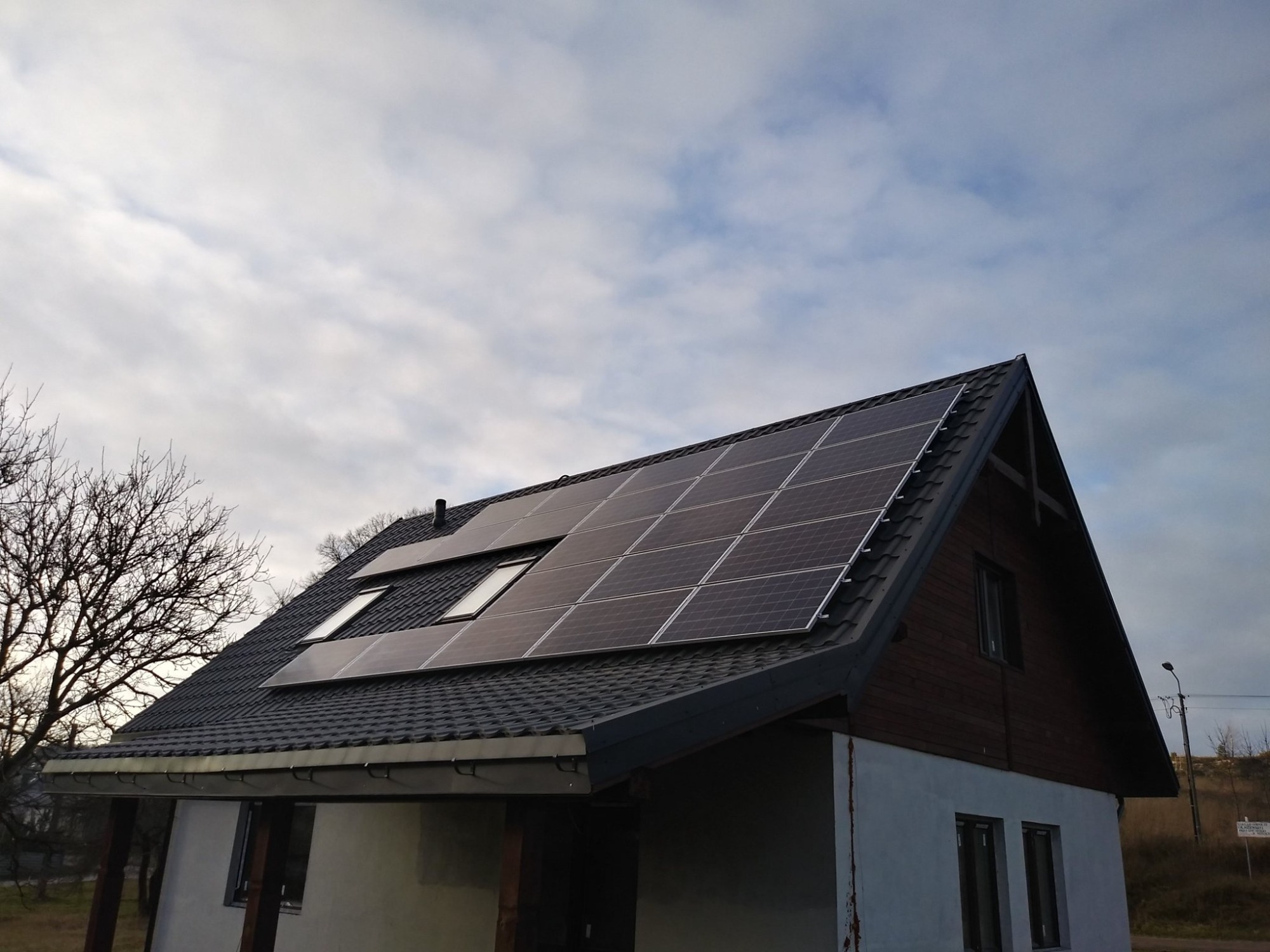 Dom jednorodzinny z panelami słonecznymi na dachu, widoczne okna dachowe, pochmurne niebo w tle.