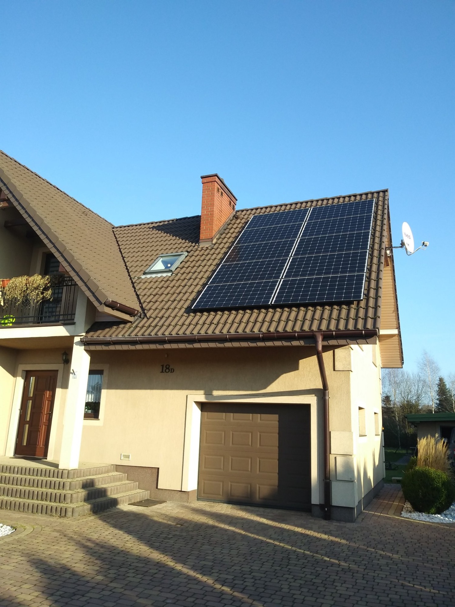 Dom jednorodzinny z brązowym dachem i panelami słonecznymi na połaci, antena satelitarna, jasna elewacja, kostka brukowa na podjeździe.