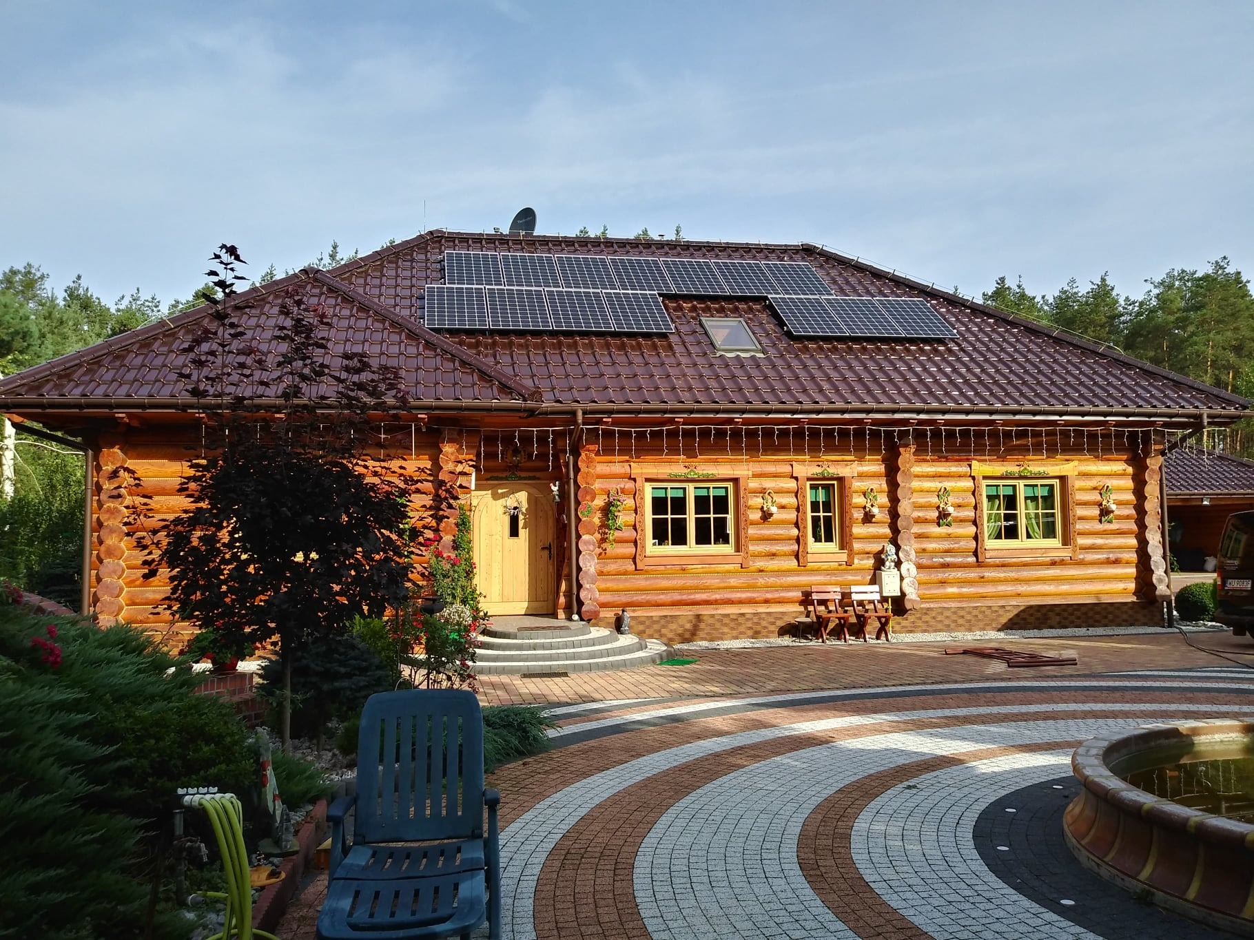 Drewniany dom z brązowym dachem, na którym zamontowano panele słoneczne; widoczny ozdobny bruk przed domem i drzewa w tle.