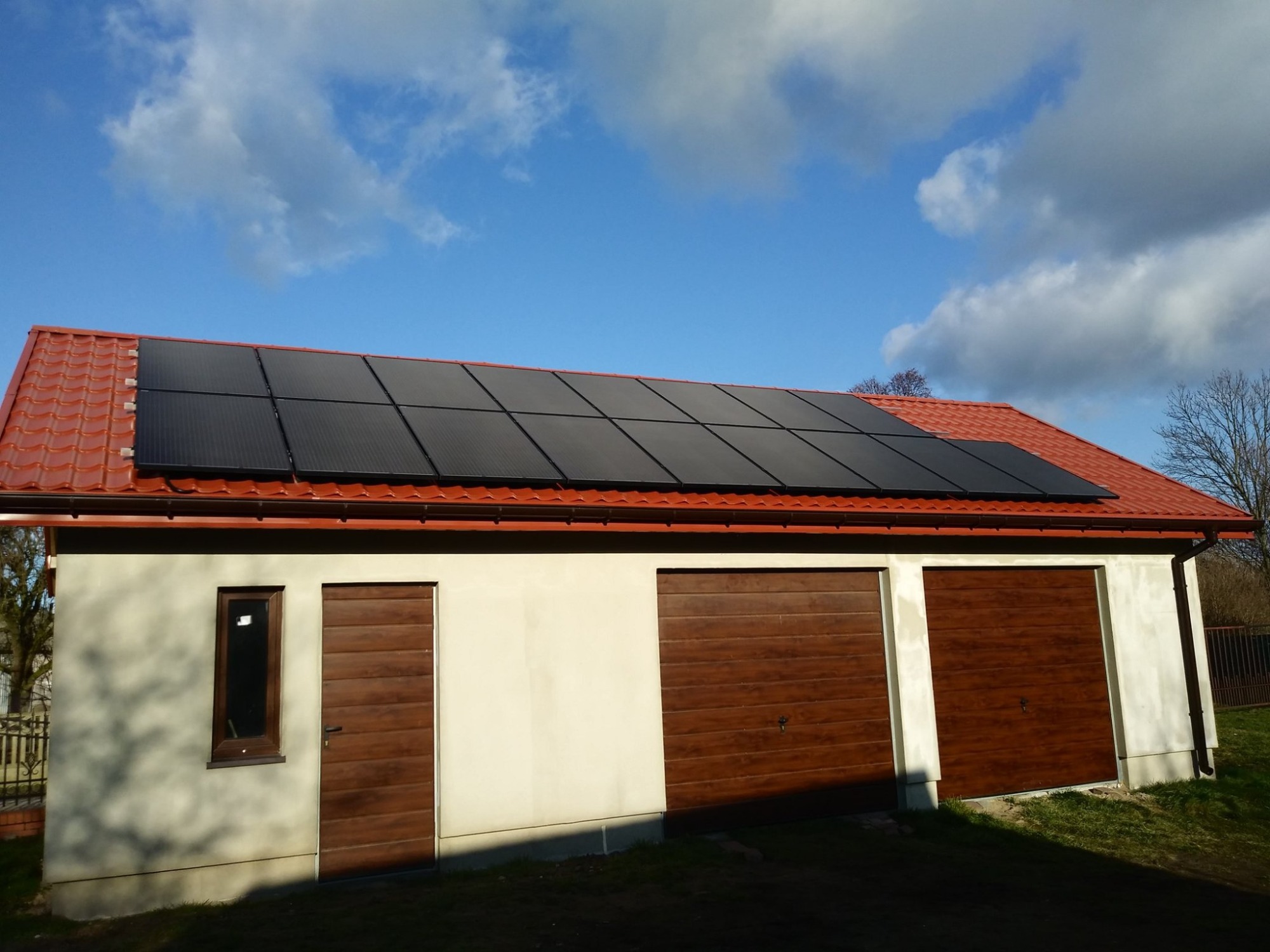 Czarny panel słoneczny na czerwonym dachu garażu z drewnianymi drzwiami garażowymi i wejściowymi pod błękitnym niebem z chmurami.