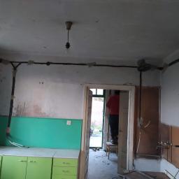ELPRO Usługi Elektryczne Dawid Olszewski - Wnętrze remontowanego pomieszczenia z widoczną instalacją elektryczną prowadzoną po ścianach, osoba na krześle w tle, wisząca żarówka i częściowo pomalowane szafki kuchenne.