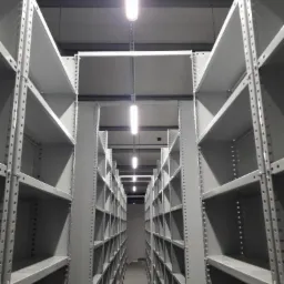 Długi korytarz między rzędami wysokich, metalowych regałów magazynowych w hali przemysłowej, oświetlony podłużnymi lampami sufitowymi.