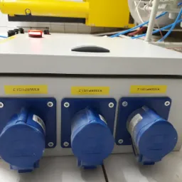 Szara skrzynka elektryczna z trzema niebieskimi gniazdami przemysłowymi, oznaczona żółtymi etykietami R3/WRP1/S1.3, R3/WRP1/S1.2 i R3/WRP1/S1.4, z widocznymi przewodami i elementami instalacji w tle.