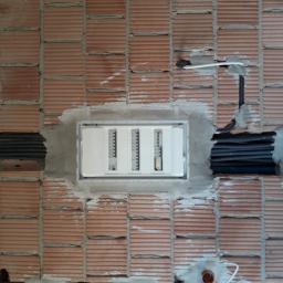 ELPRO Usługi Elektryczne Dawid Olszewski - Biała skrzynka rozdzielcza z bezpiecznikami zamontowana w surowej, ceglanej ścianie z widocznymi kablami w peszlach i świeżym cementem.
