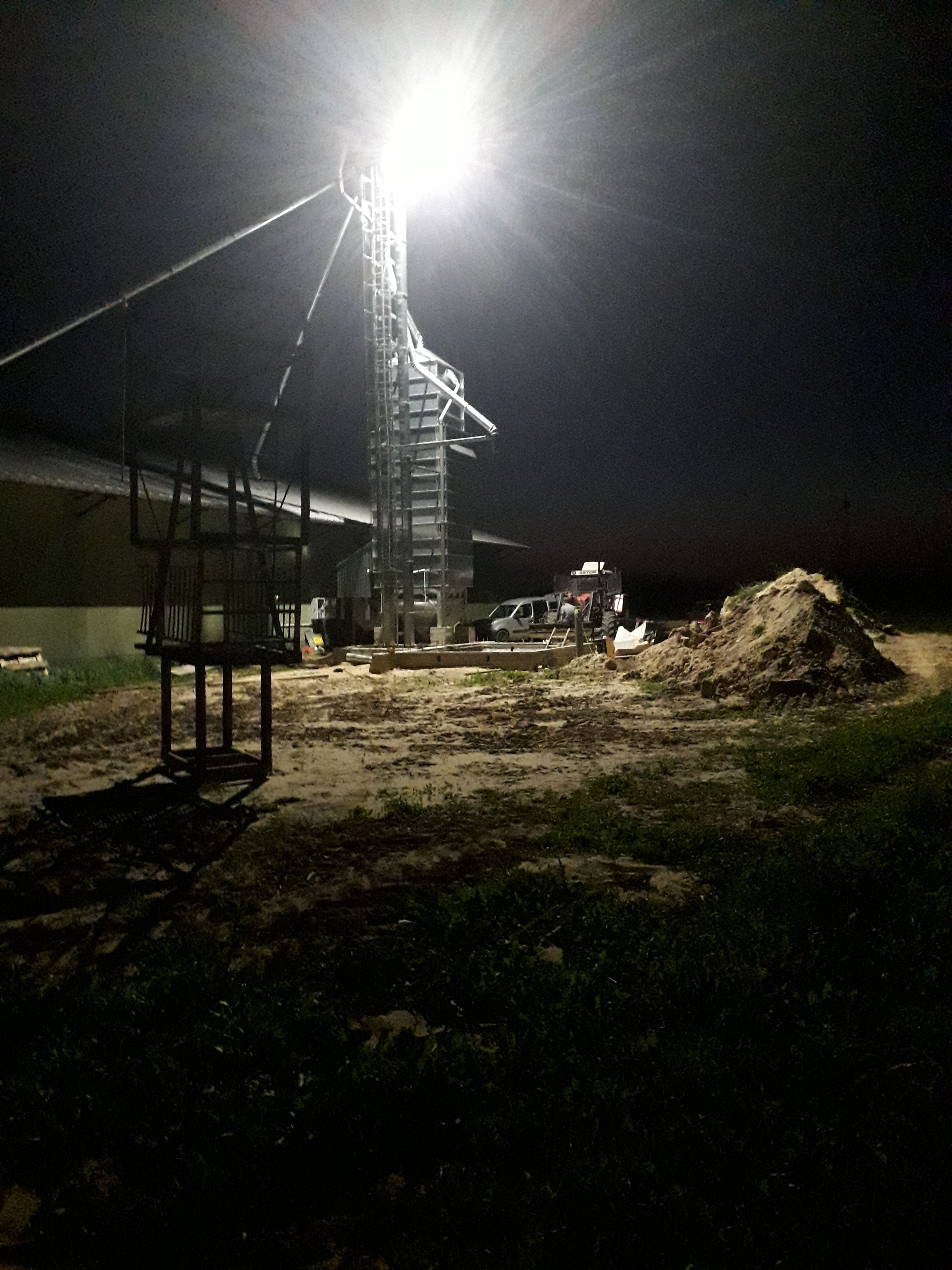 Nocna fotografia budowy z metalową konstrukcją oświetloną jasnym światłem, widoczny sprzęt budowlany, wykopana ziemia i częściowo zarys budynku w tle.