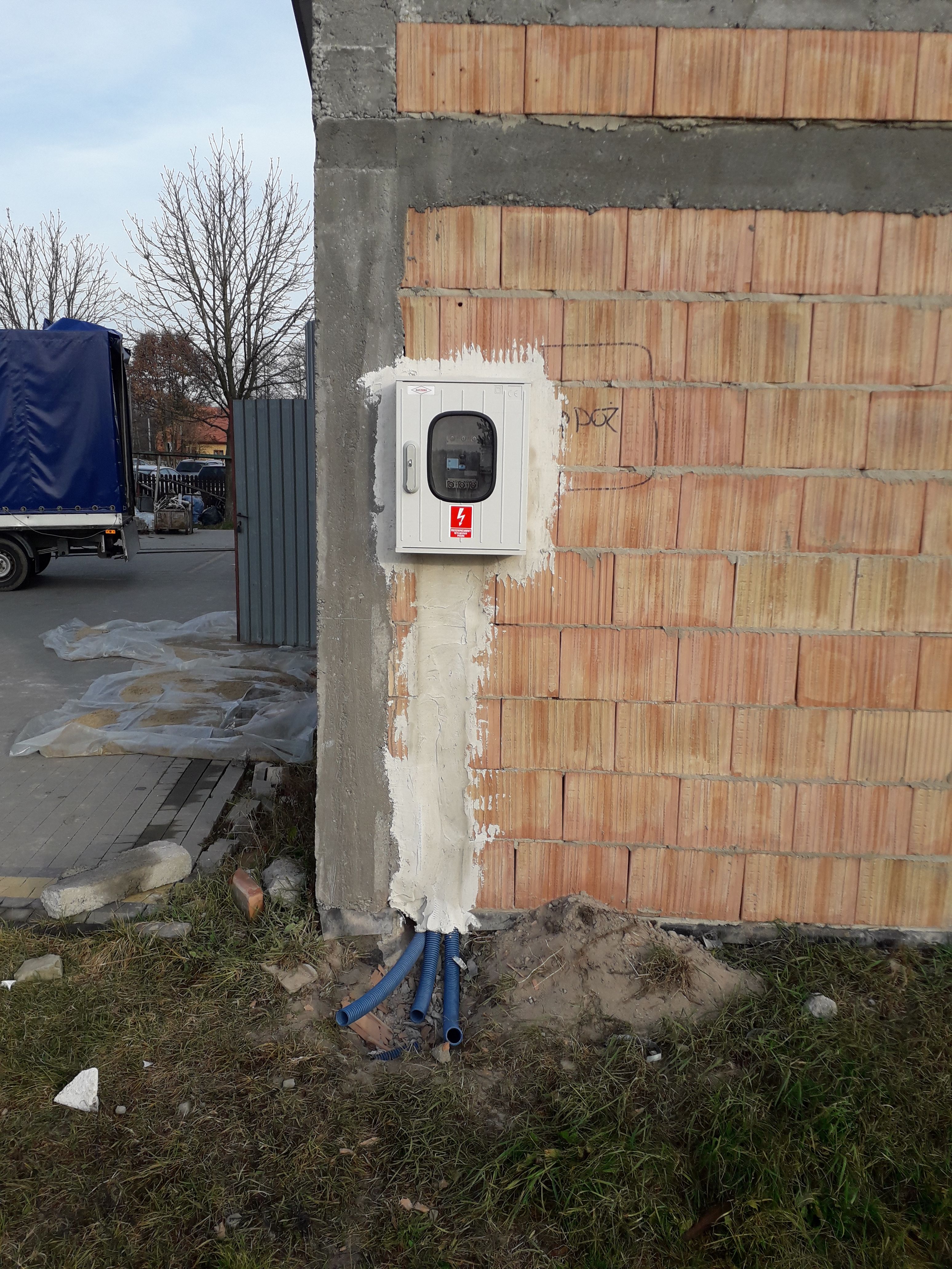 Szara skrzynka elektryczna z oznaczeniem ostrzegawczym przymocowana do ściany z czerwonej cegły, z widocznymi niebieskimi rurami instalacyjnymi wychodzącymi z dołu, świeżo obrobiona zaprawą,...