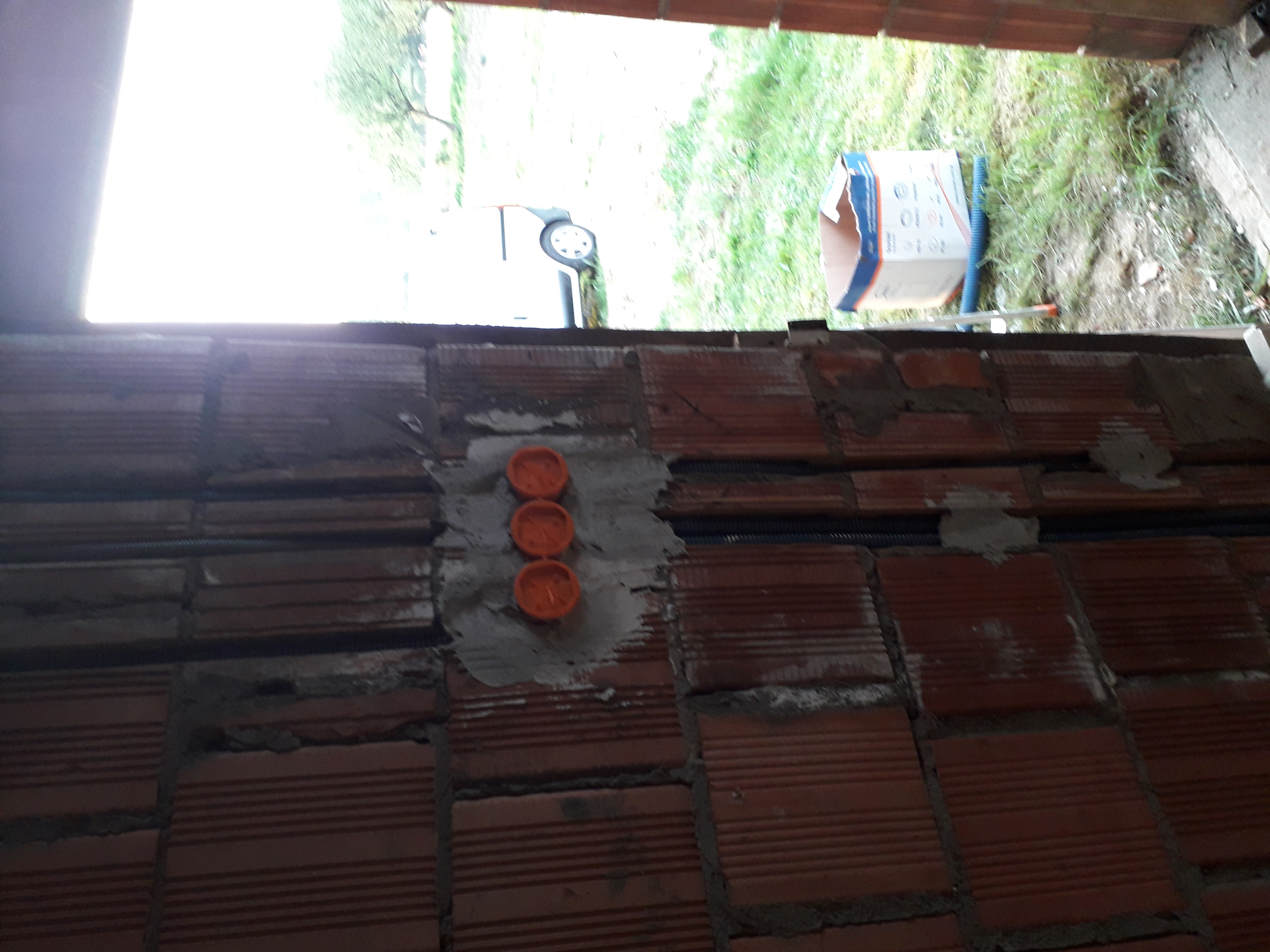 Fragment ściany z cegły dziurawki z trzema pomarańczowymi puszkami elektrycznymi zamontowanymi w szarym cemencie, widoczne peszle instalacyjne, w tle trawa i samochód