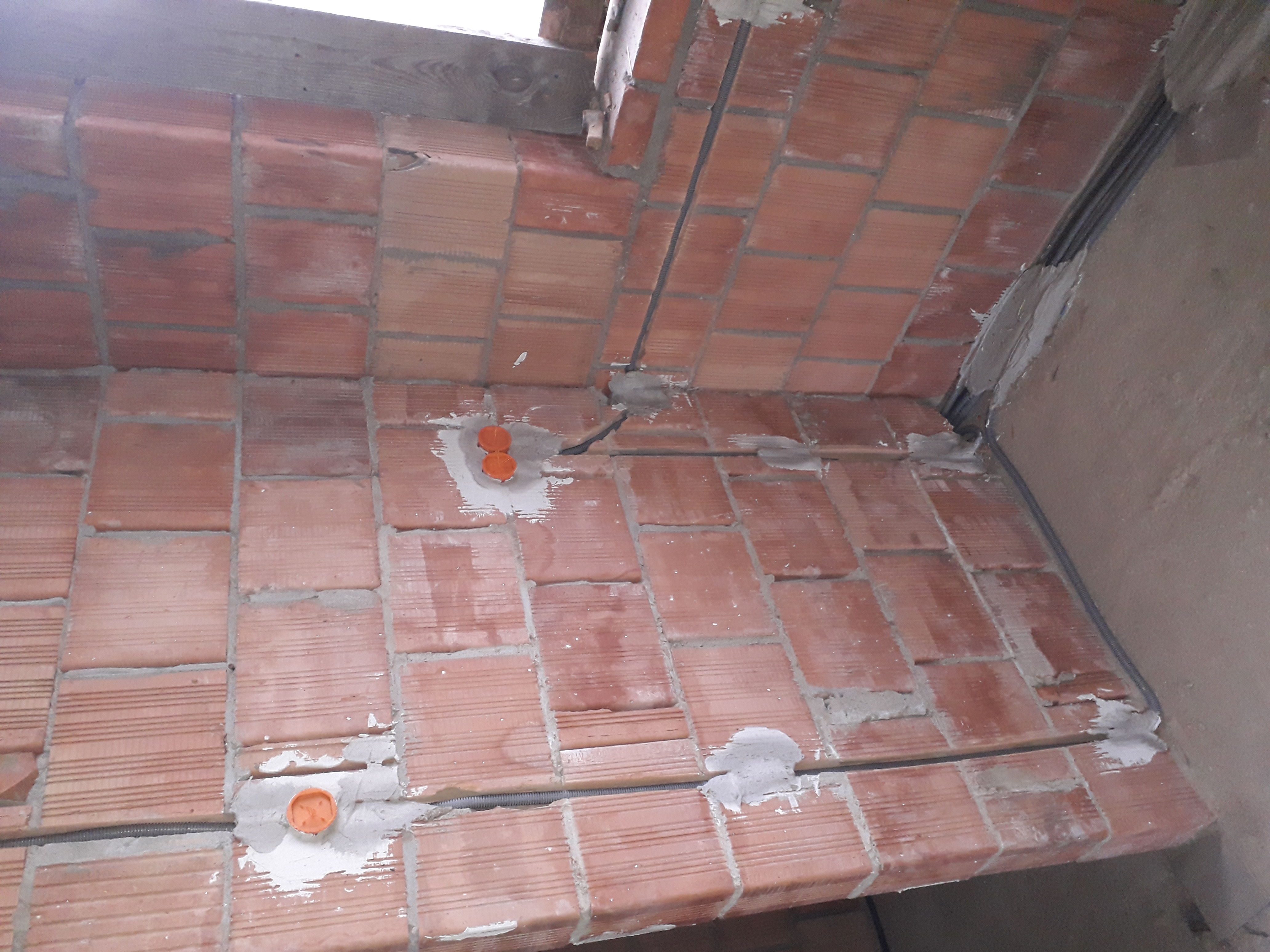 Czerwona ściana z cegieł z widocznymi przewodami elektrycznymi w peszlach i pomarańczowymi puszkami instalacyjnymi, częściowo zamurowanymi.