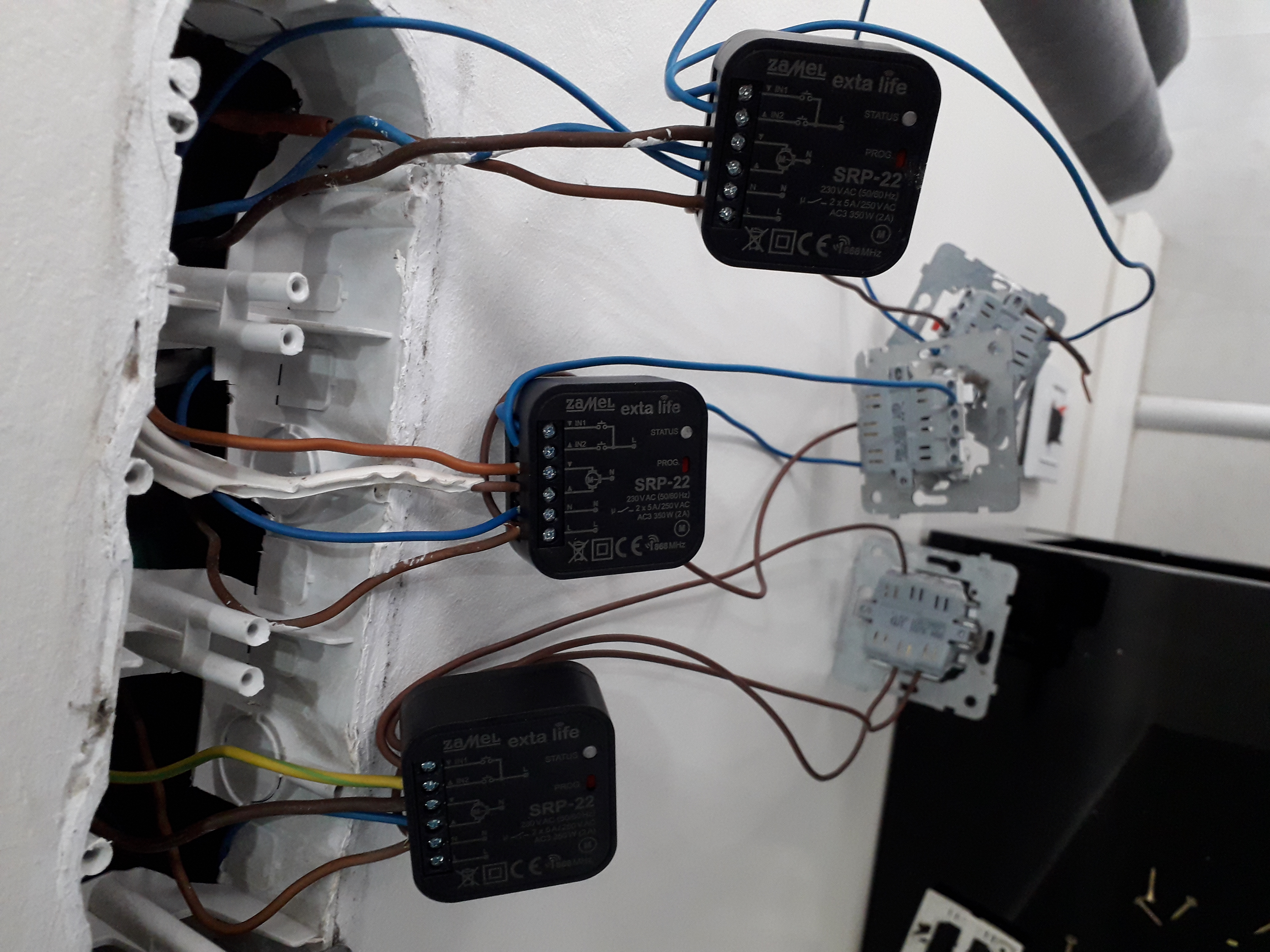 Fragment instalacji elektrycznej w trakcie montażu, widoczne trzy moduły sterujące Zamel Exta Life SRP-22 podłączone przewodami do puszek instalacyjnych i gniazd w ścianie, z bliska, z widocznymi...