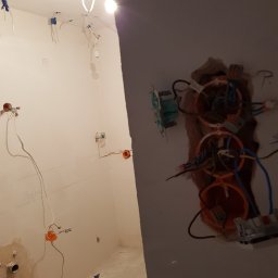 Tomasz Graczyk - Wnętrze w trakcie remontu: widoczne przewody elektryczne wystające ze ściany i sufitu, puszki instalacyjne w kolorze pomarańczowym, wisząca żarówka oświetlająca pomieszczenie, ślady po instalacjach...