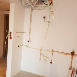 Tomasz Graczyk - Ściana w trakcie prac instalacyjnych: widoczne przewody elektryczne, puszki podtynkowe, rury wentylacyjne pod sufitem oraz narzędzia malarskie oparte o ścianę.