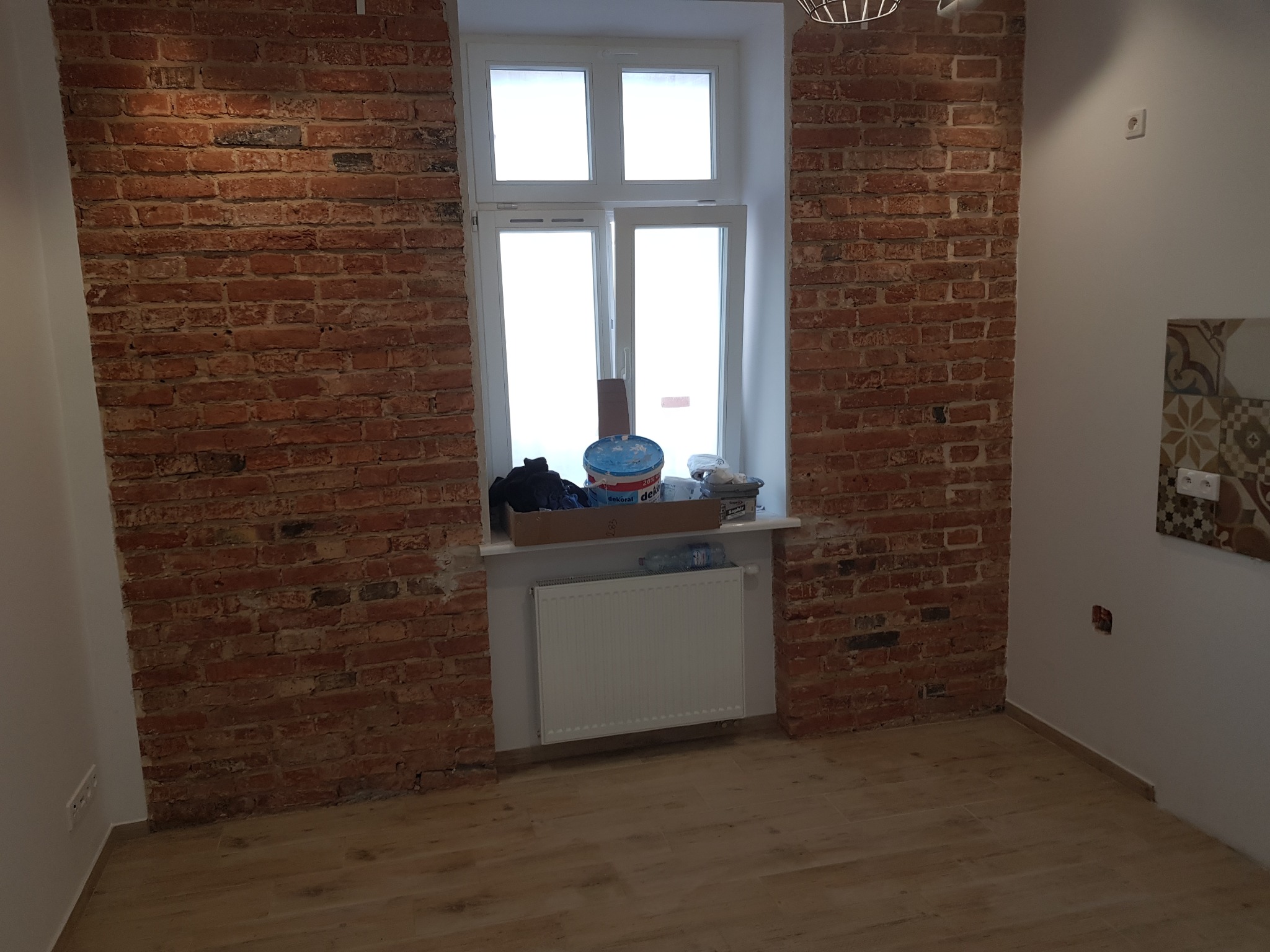 Pokój z odsłoniętą czerwoną cegłą na ścianach, białe okno z farbami i narzędziami na parapecie, drewniana podłoga w trakcie prac wykończeniowych.