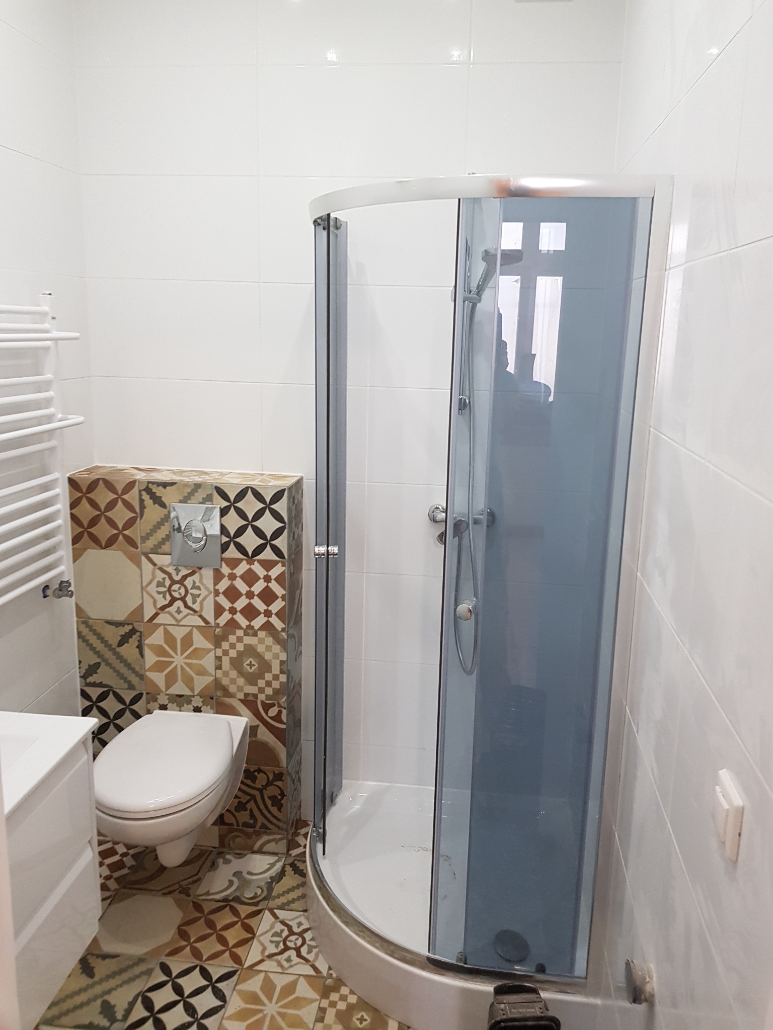 Kompaktowa łazienka z kabiną prysznicową z szarymi szybami, toaletą podwieszaną obudowaną mozaiką i grzejnikiem drabinkowym na tle białych płytek.