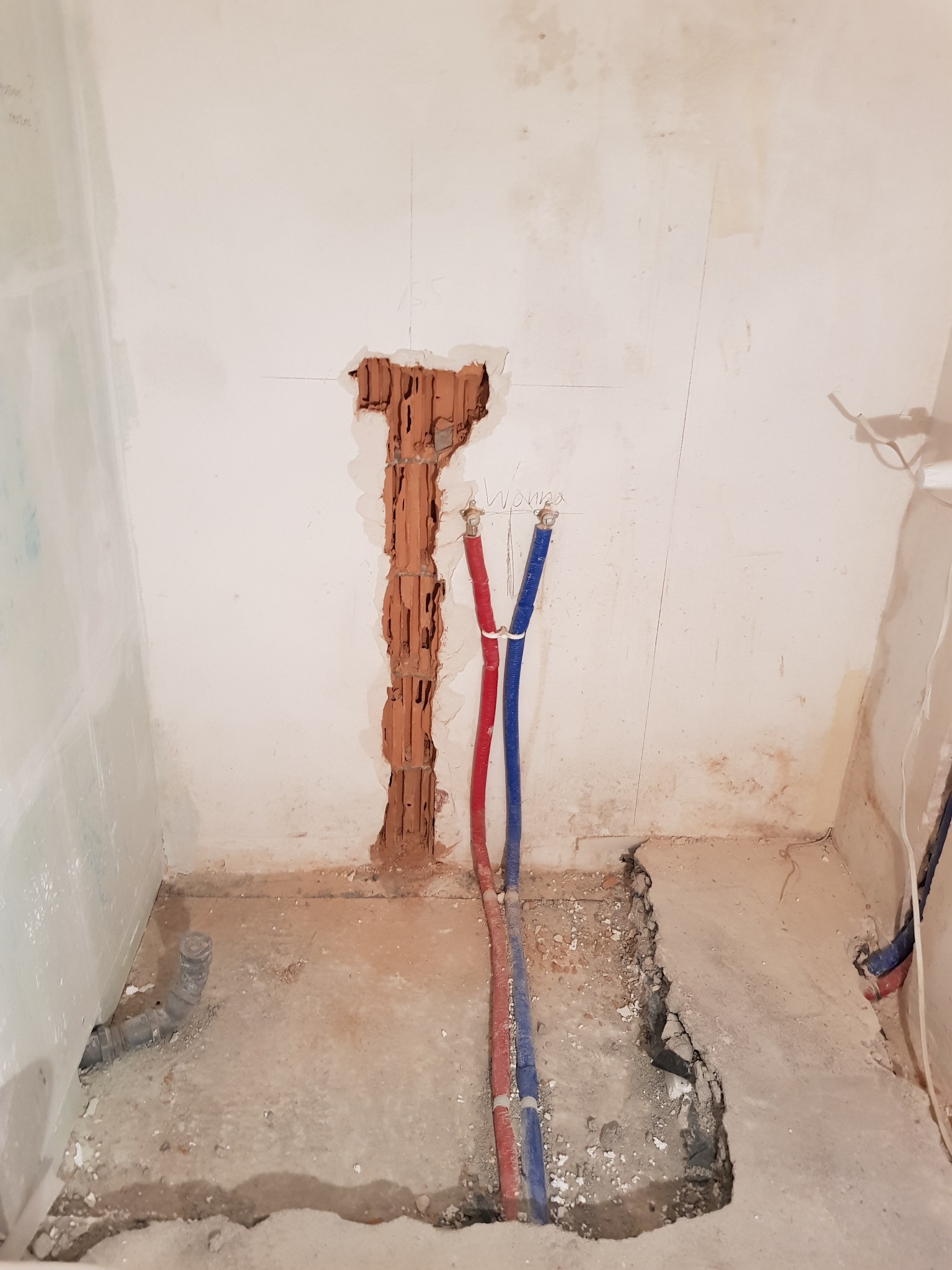 Przygotowanie do instalacji wodnej w łazience: widoczne wystające rury (czerwona i niebieska) oraz fragment ściany z odsłoniętą cegłą, pozostałości po kuciu w posadzce.