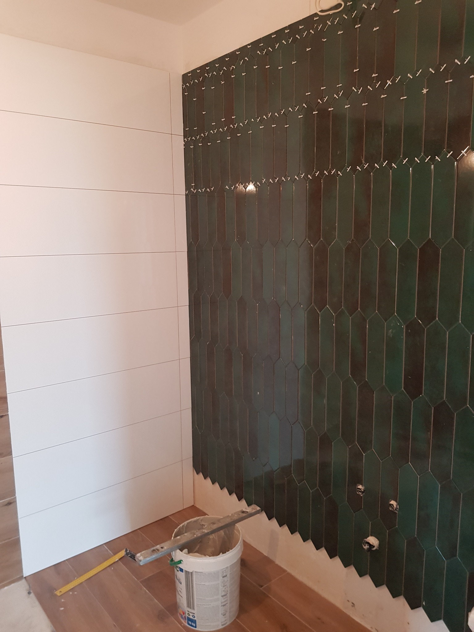 Układanie ciemnozielonych, podłużnych płytek w kształcie rombów na ścianie łazienki, widoczne krzyżyki dystansowe, obok białe płytki w poziomie, na podłodze brązowe panele, wiadro z zaprawą...