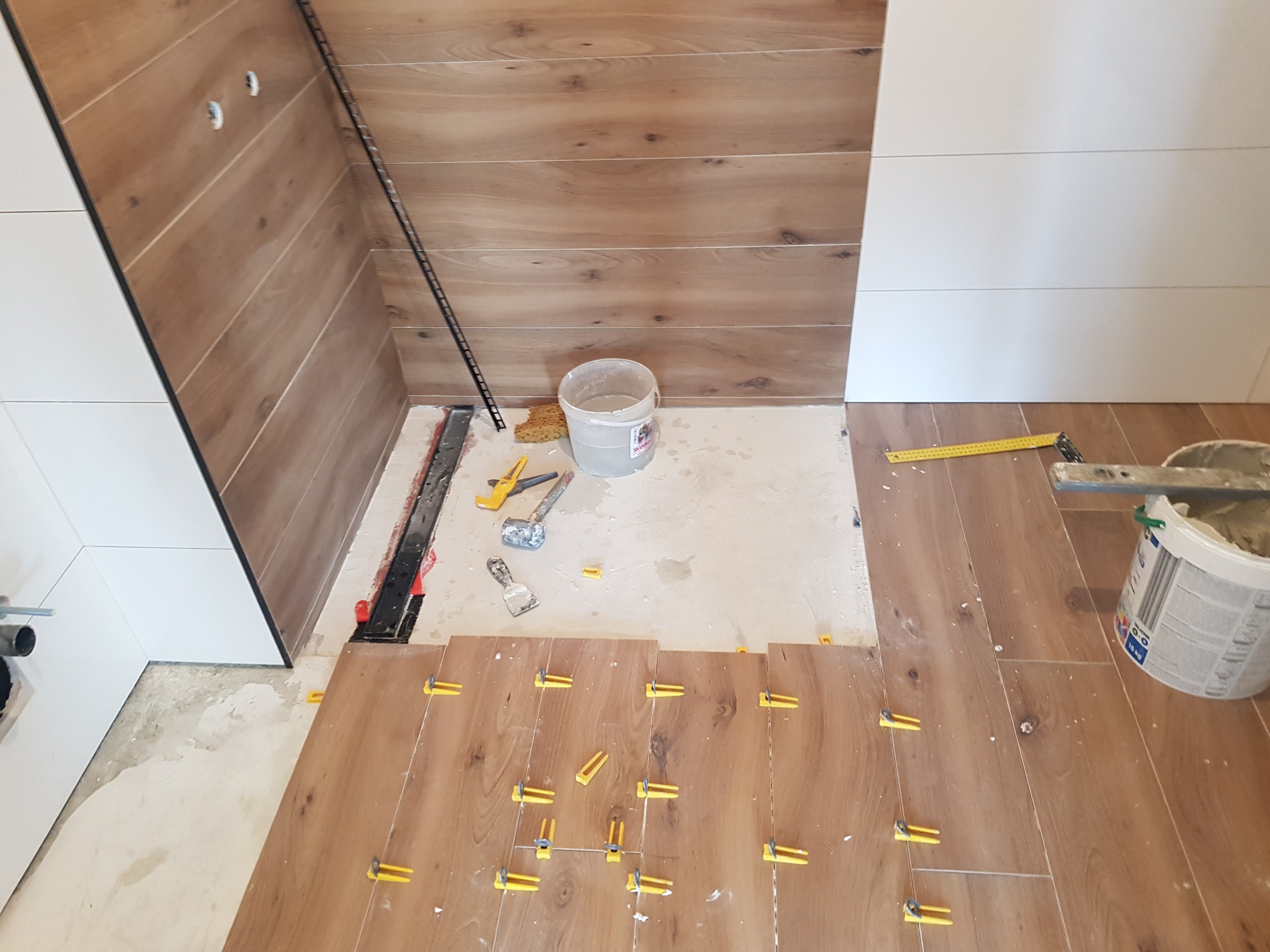 Układanie płytek imitujących drewno w łazience z użyciem klipsów poziomujących, widoczny kubeł z klejem i narzędzia glazurnicze.