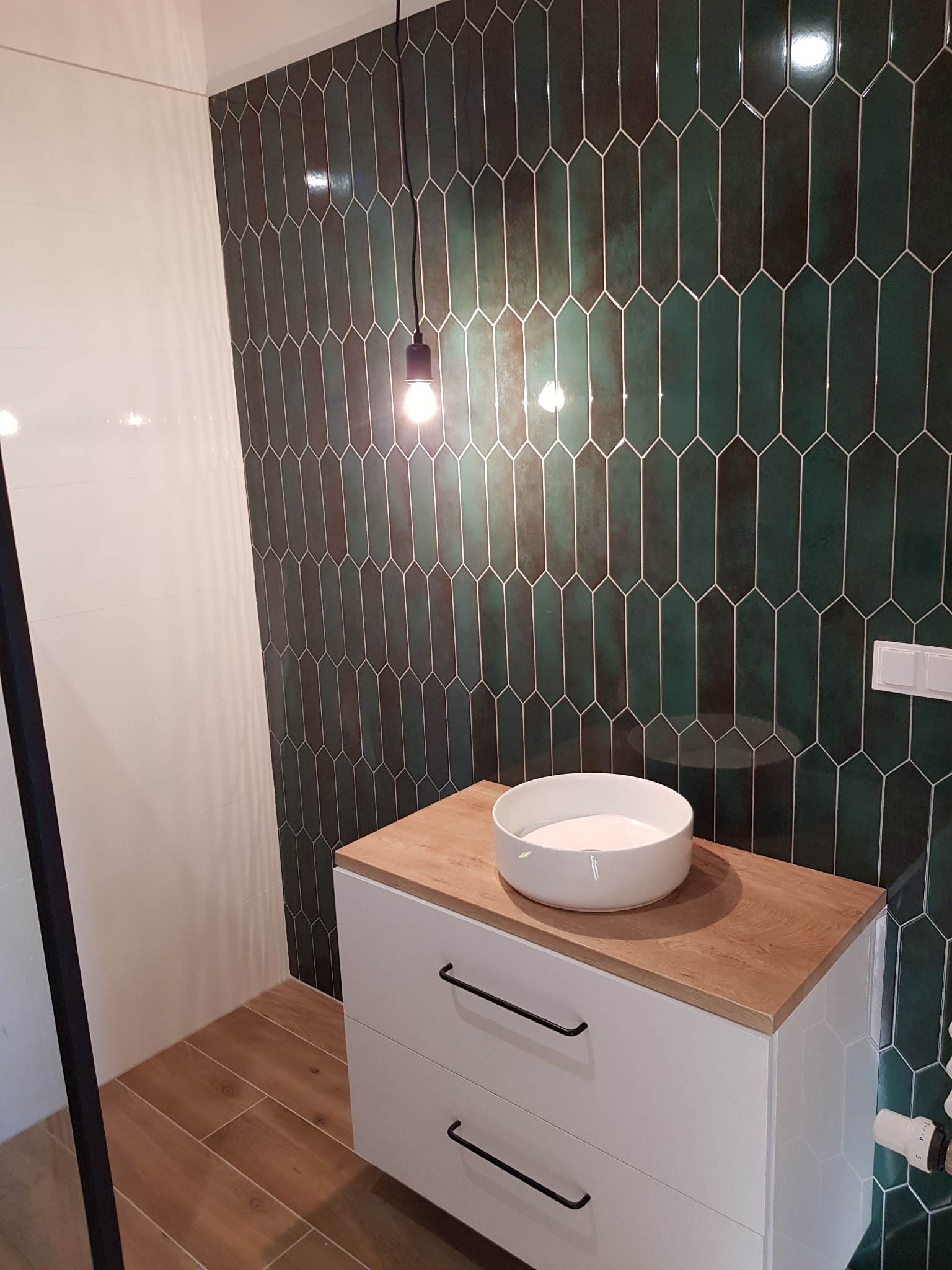 Nowoczesna łazienka z umywalką nablatową, szafką z białymi frontami i drewnianym blatem, oraz zielonymi, podłużnymi płytkami na ścianie i drewnopodobną podłogą.