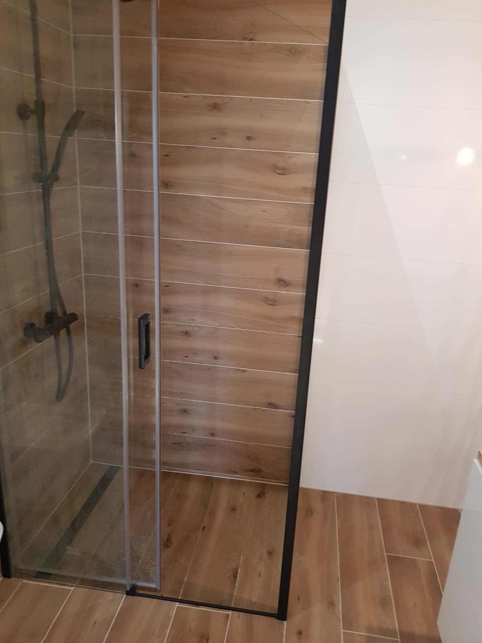 Kabina prysznicowa z czarnymi profilami, wyłożona płytkami imitującymi drewno, z widoczną baterią prysznicową w ciemnym kolorze i fragmentem białej ściany.