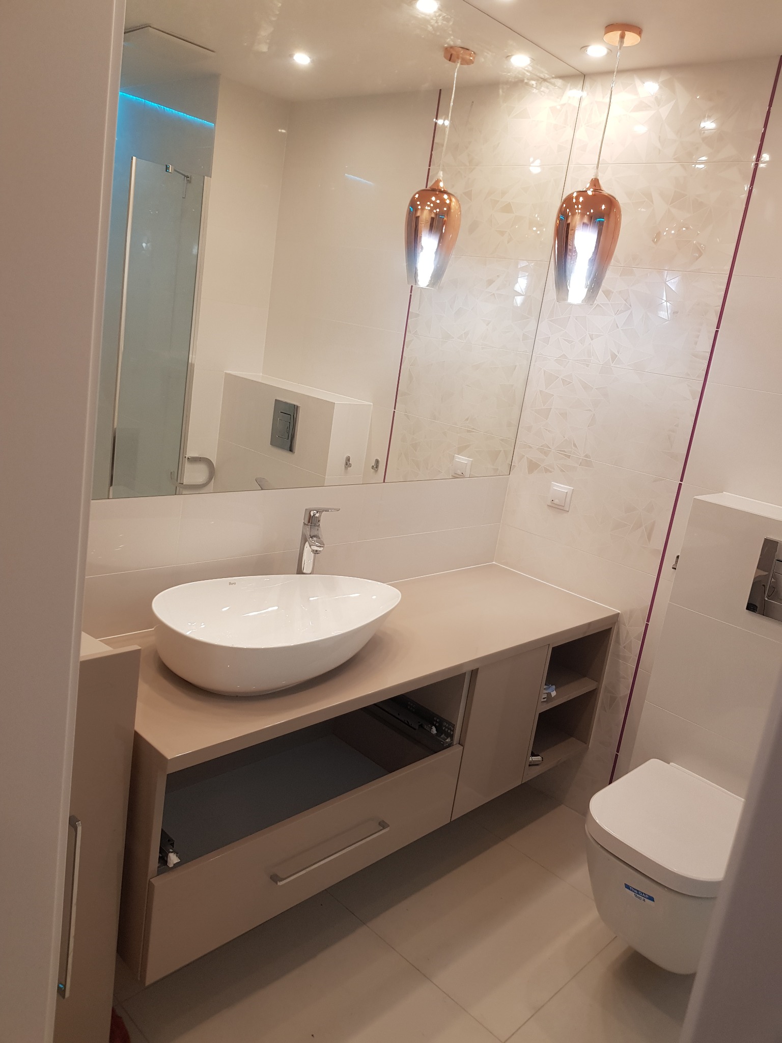 Nowoczesna łazienka z owalną umywalką nablatową, beżową szafką pod umywalką z otwartymi półkami, wiszącą toaletą i dekoracyjnymi lampami wiszącymi w kolorze miedzi.