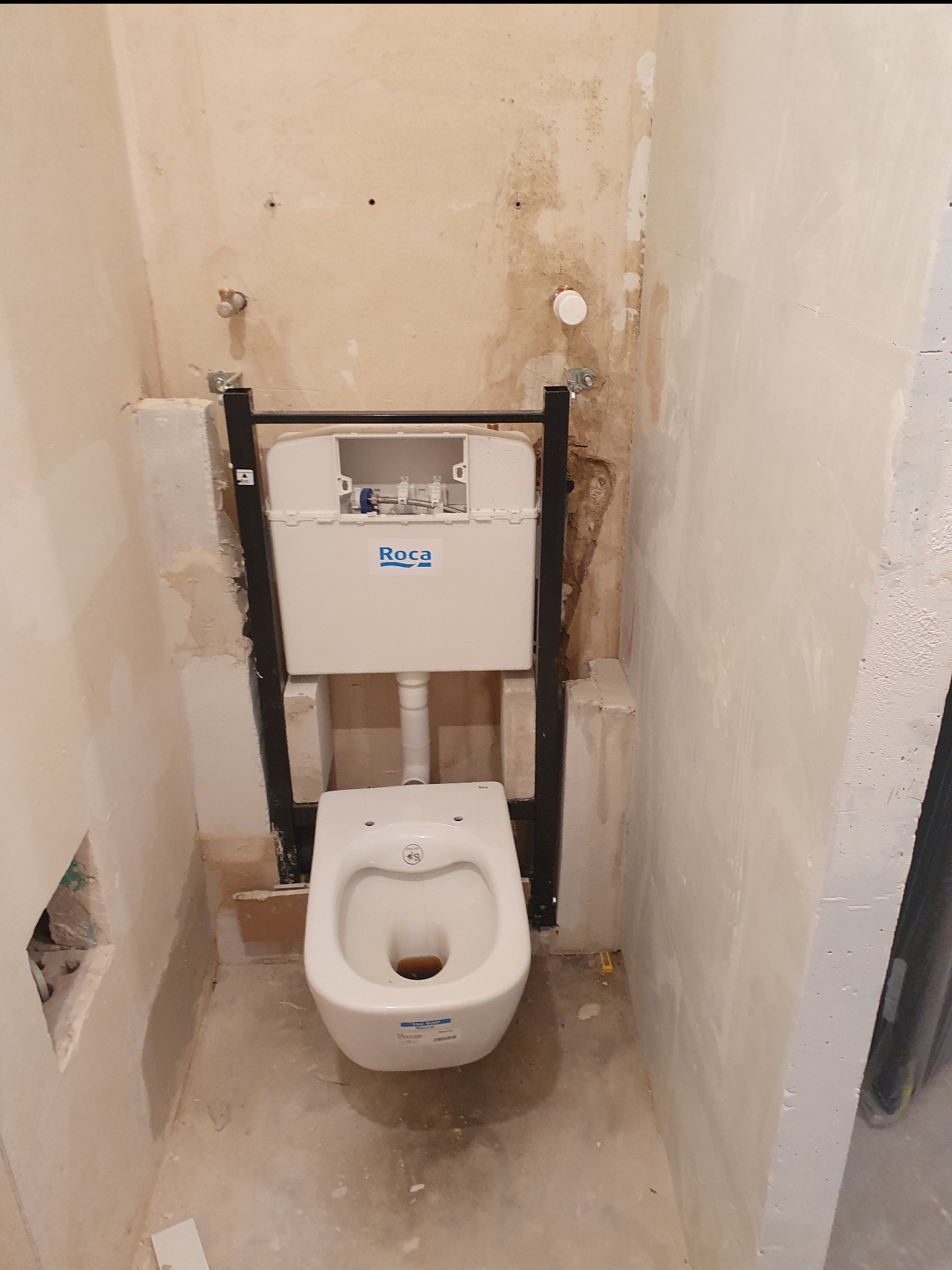 Stelaż podtynkowy z zamontowaną białą miską WC Roca w surowym stanie łazienki przed wykończeniem, widoczne ślady po kuciu w ścianach.