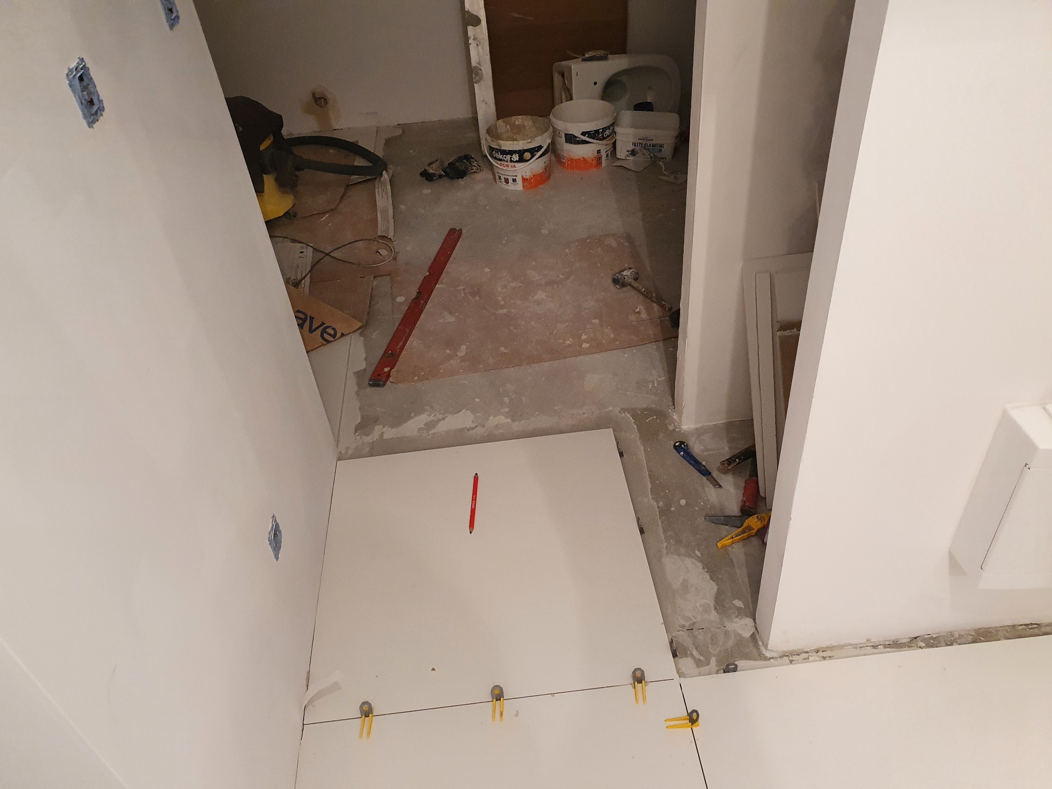 Układanie białych płytek podłogowych w łazience, widoczne krzyżyki dystansowe, poziomica, ołówek na płytce, puszki z farbą w tle.