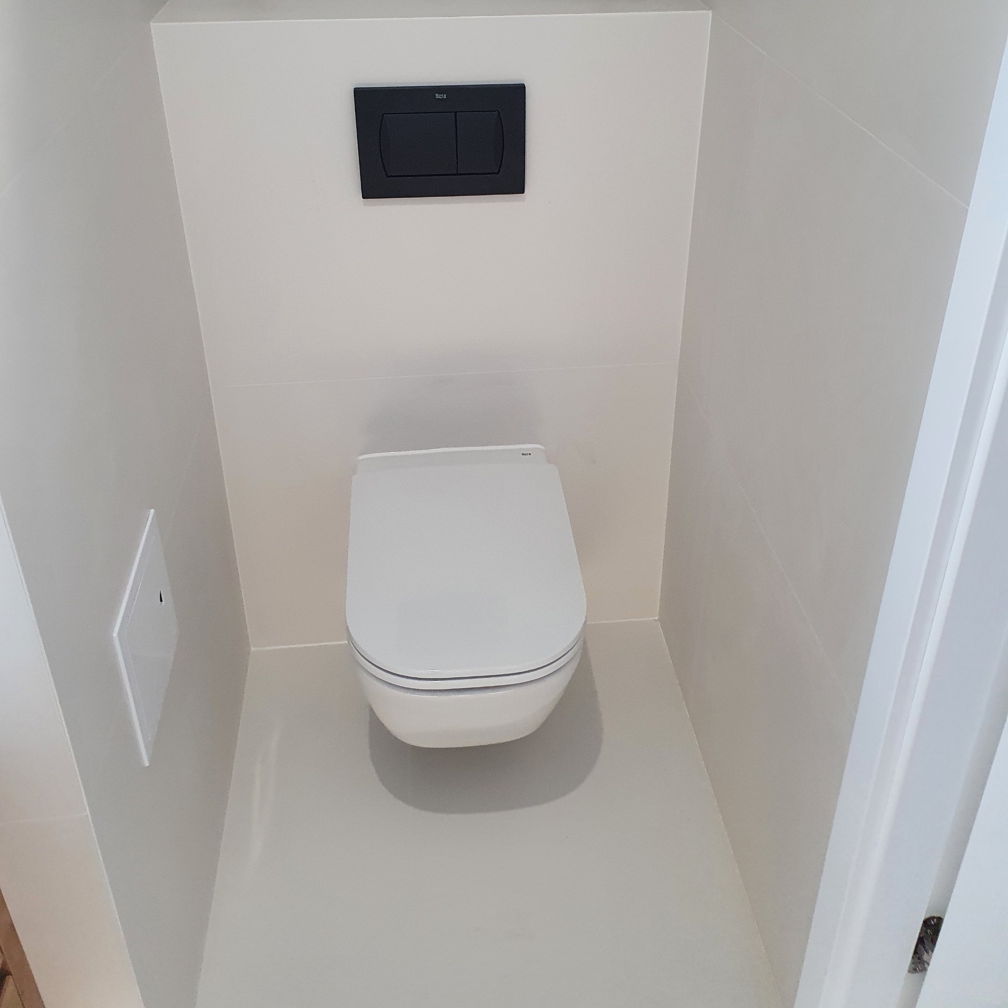 Wisząca miska WC z czarnym przyciskiem spłukującym w minimalistycznej łazience z jasnymi płytkami.