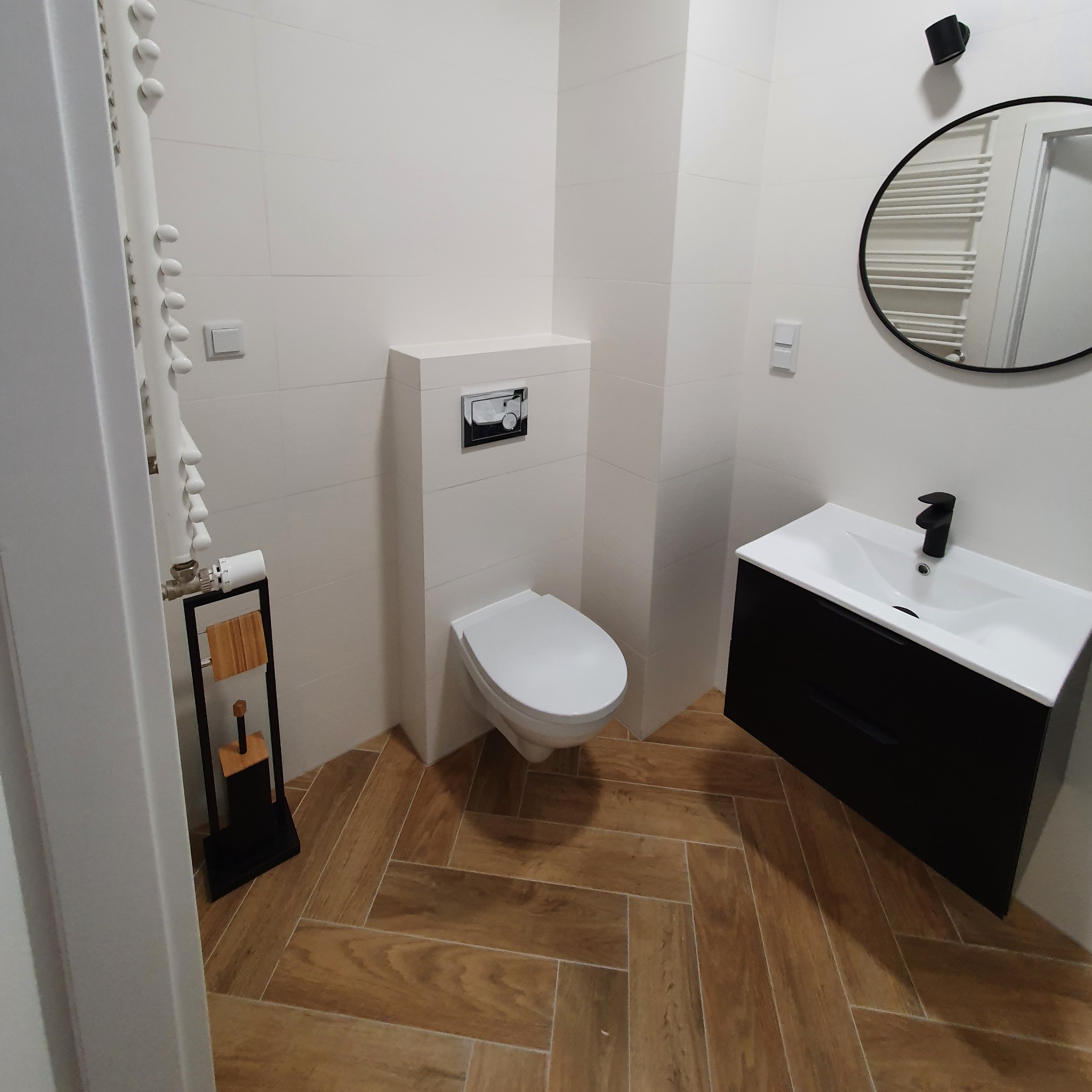 Nowoczesna łazienka z białymi płytkami ściennymi, drewnopodobną podłogą w jodełkę, wiszącą toaletą, czarną szafką pod umywalką z białą umywalką i okrągłym lustrem w czarnej ramie.