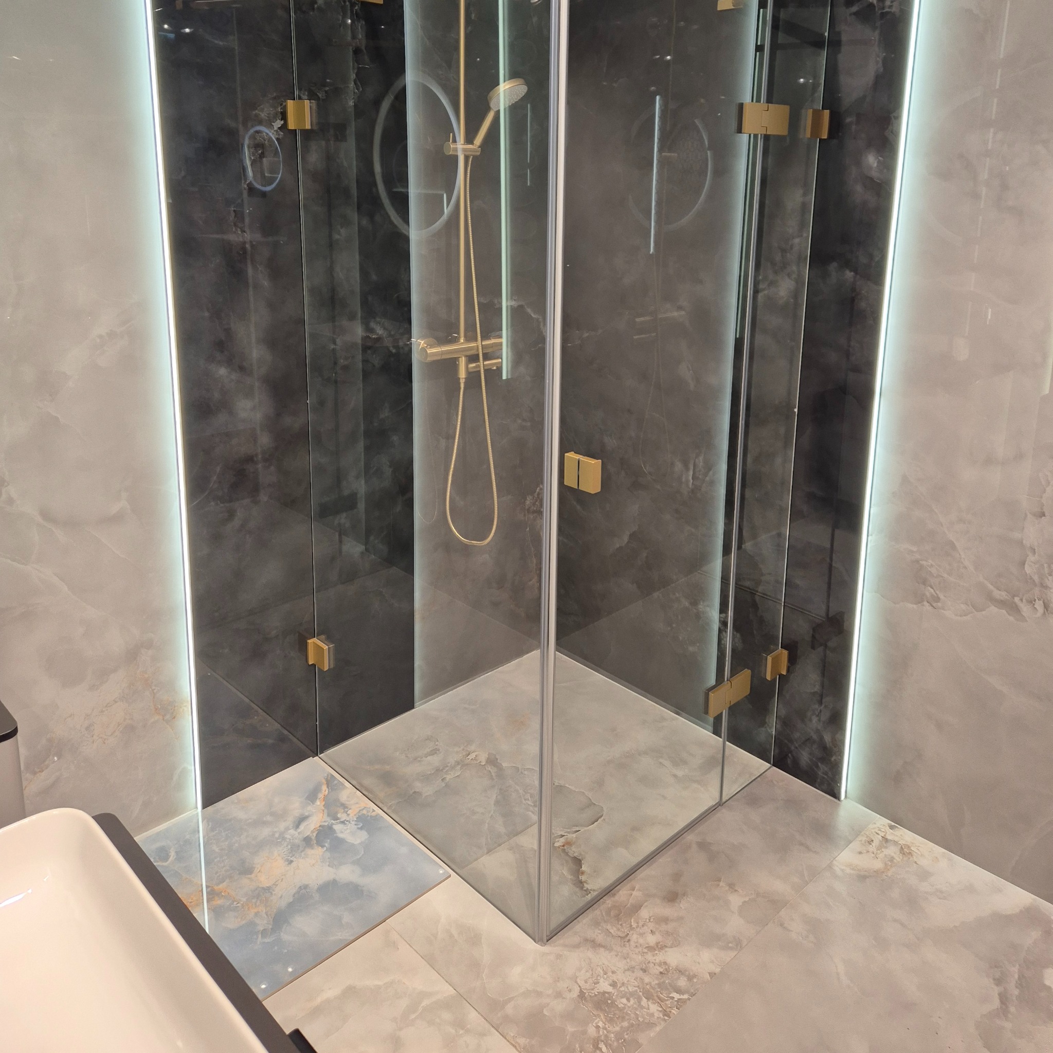 Szklana kabina prysznicowa z deszczownicą w kolorze złotym, zamontowana w rogu łazienki wyłożonej płytkami imitującymi marmur w odcieniach szarości i beżu, z oświetleniem LED wzdłuż krawędzi ściany.