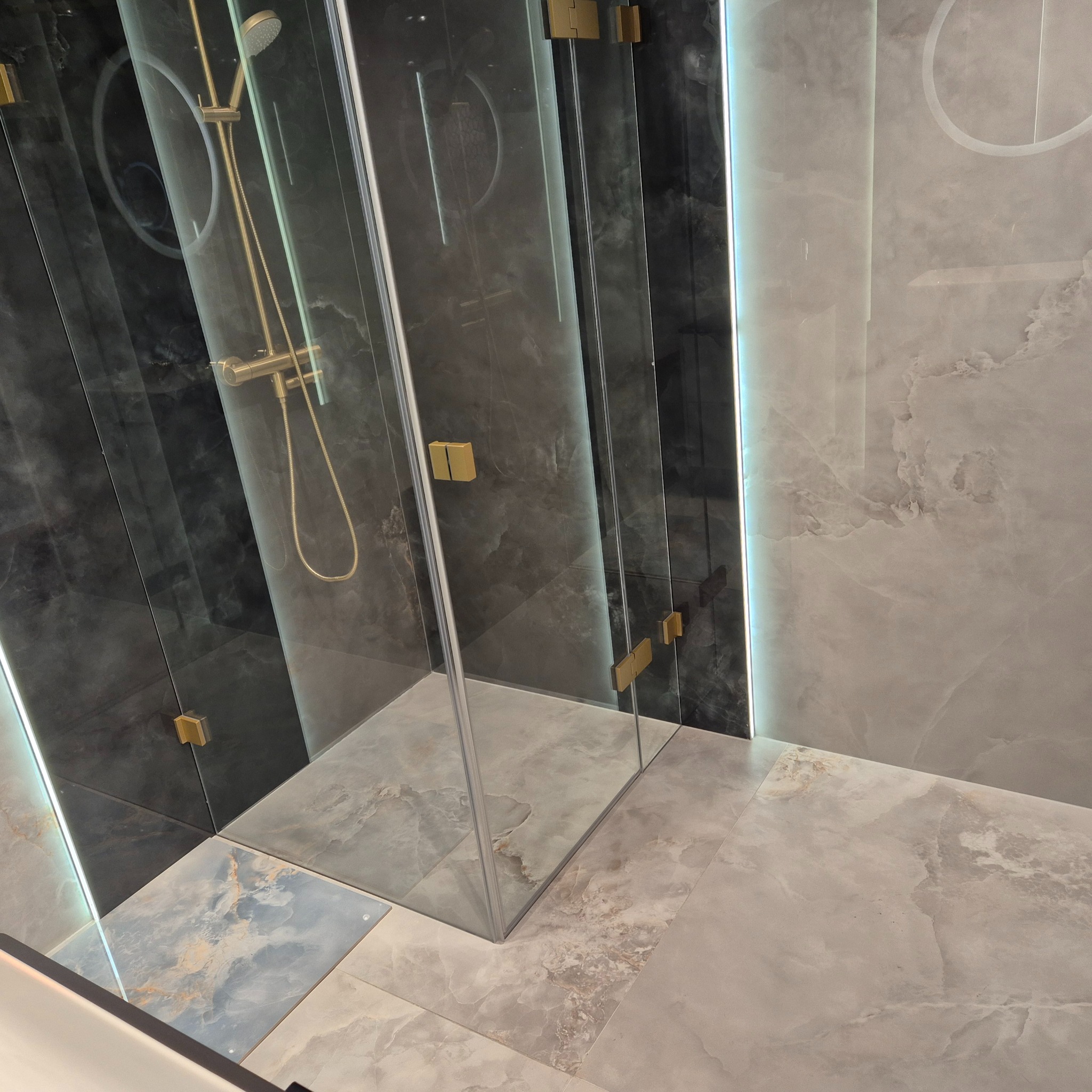 Nowoczesna kabina prysznicowa z złotą armaturą, szklane drzwi z minimalistycznymi zawiasami, marmurowe płytki w odcieniach szarości i czerni, podświetlenie LED na ścianie.