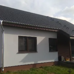 Gotowy, parterowy budynek z ciemną dachówką, brązowymi ramami okiennymi i szarymi ścianami, z widocznymi rynnami i częściowo wykończonym tarasem z kostki.