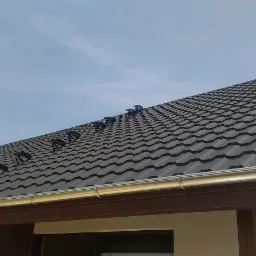 Ciemnoszara blachodachówka z systemem odśnieżania i rynnami, widok z dołu na fragment domu pod błękitnym niebem.