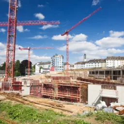Rozległy plac budowy z czerwonymi żurawiami na tle błękitnego nieba i zabudowy miejskiej. Widoczne szalunki i zbrojenia fundamentów. Prace w toku.
