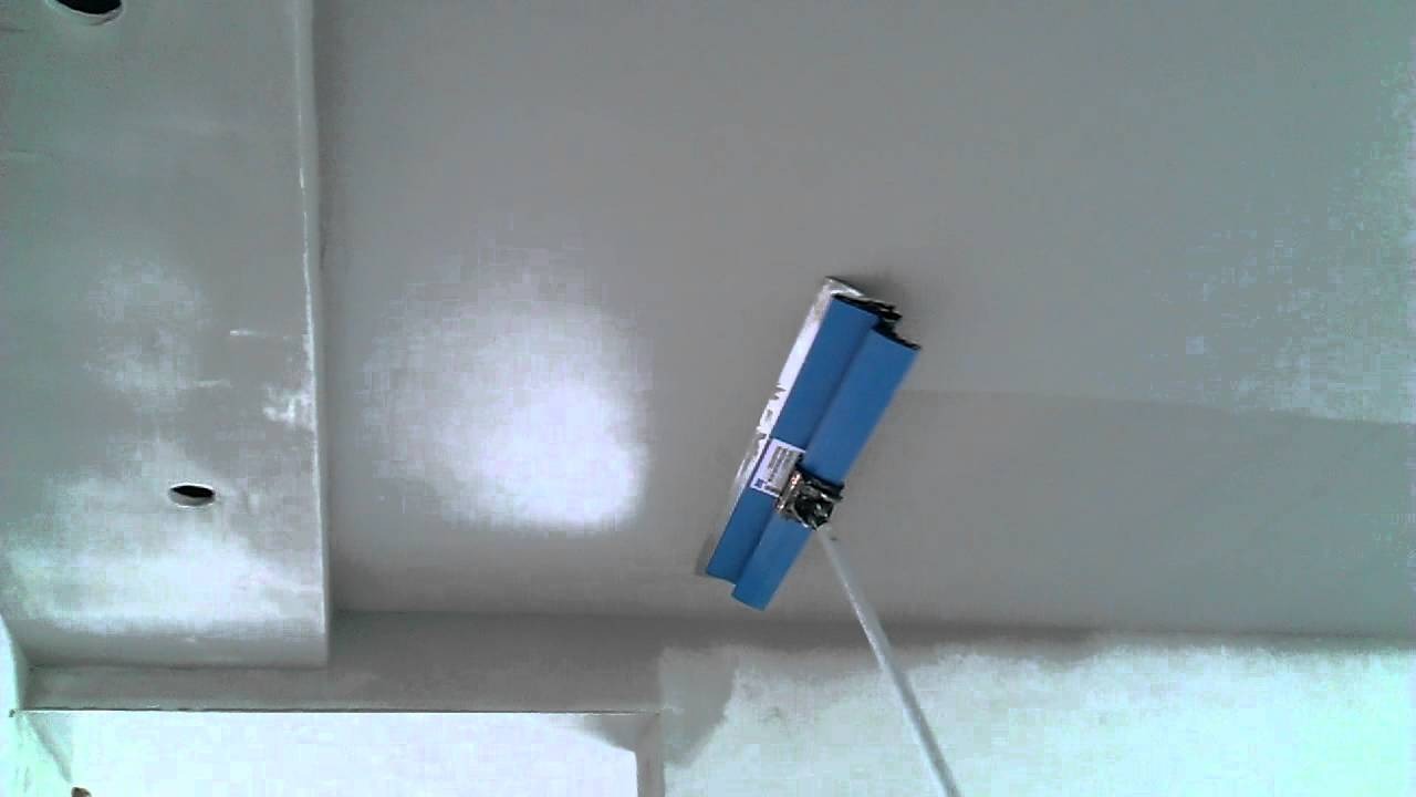 Aplikator teleskopowy z niebieskimi wałkami podczas równomiernego rozprowadzania świeżej warstwy szpachli na suficie podwieszanym z wycięciami na oświetlenie.