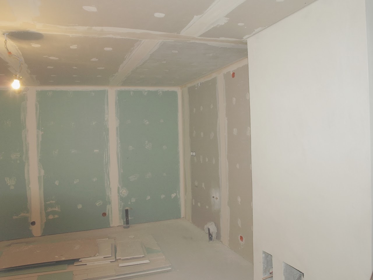 Wnętrze w trakcie prac wykończeniowych: ściany z płyt gipsowo-kartonowych z widocznymi spoinami i otworami, sufit z częściowo nałożoną gładzią, oraz stos płyt gipsowych na podłodze.