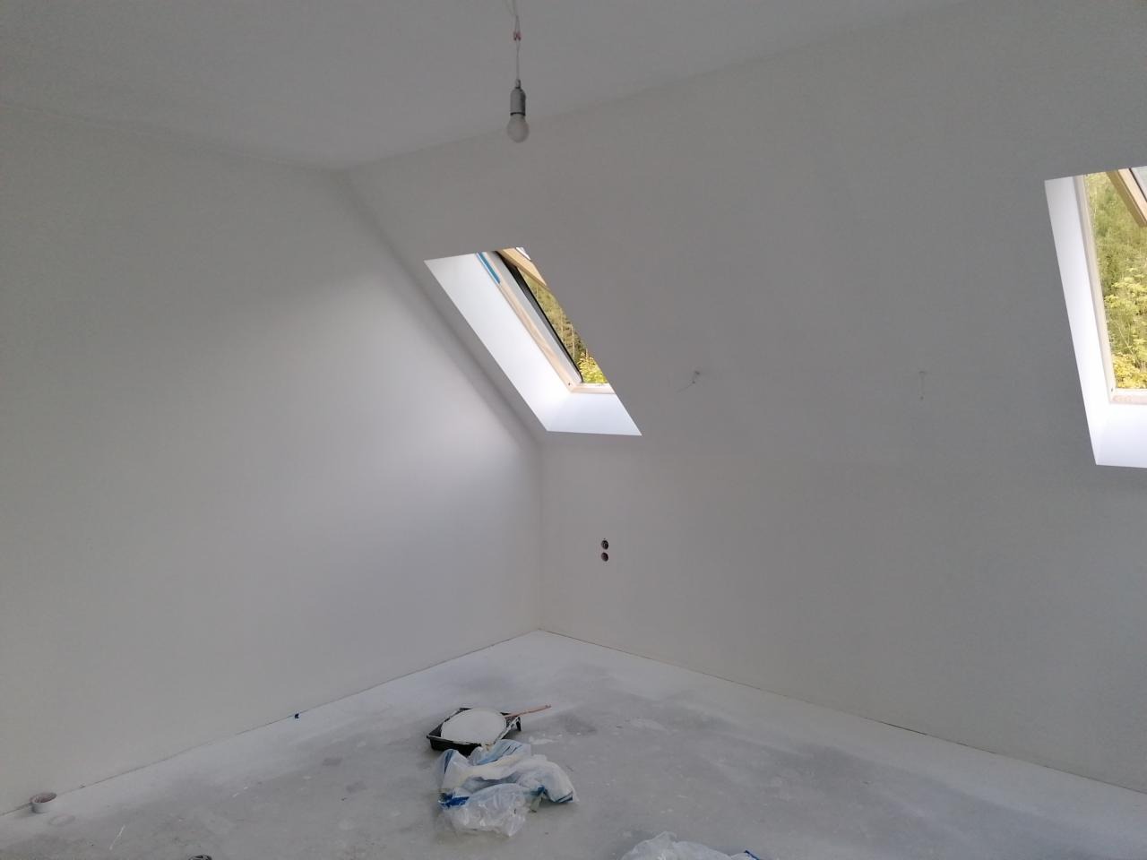Poddasze po pracach wykończeniowych, świeżo pomalowane na biało, widoczne dwa okna dachowe, na podłodze folia malarska i kuweta z farbą.