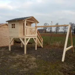 Surowa drewniana konstrukcja placu zabaw: domek na palach z balustradą, schodami i huśtawką, widok z boku na tle pola i zabudowań.