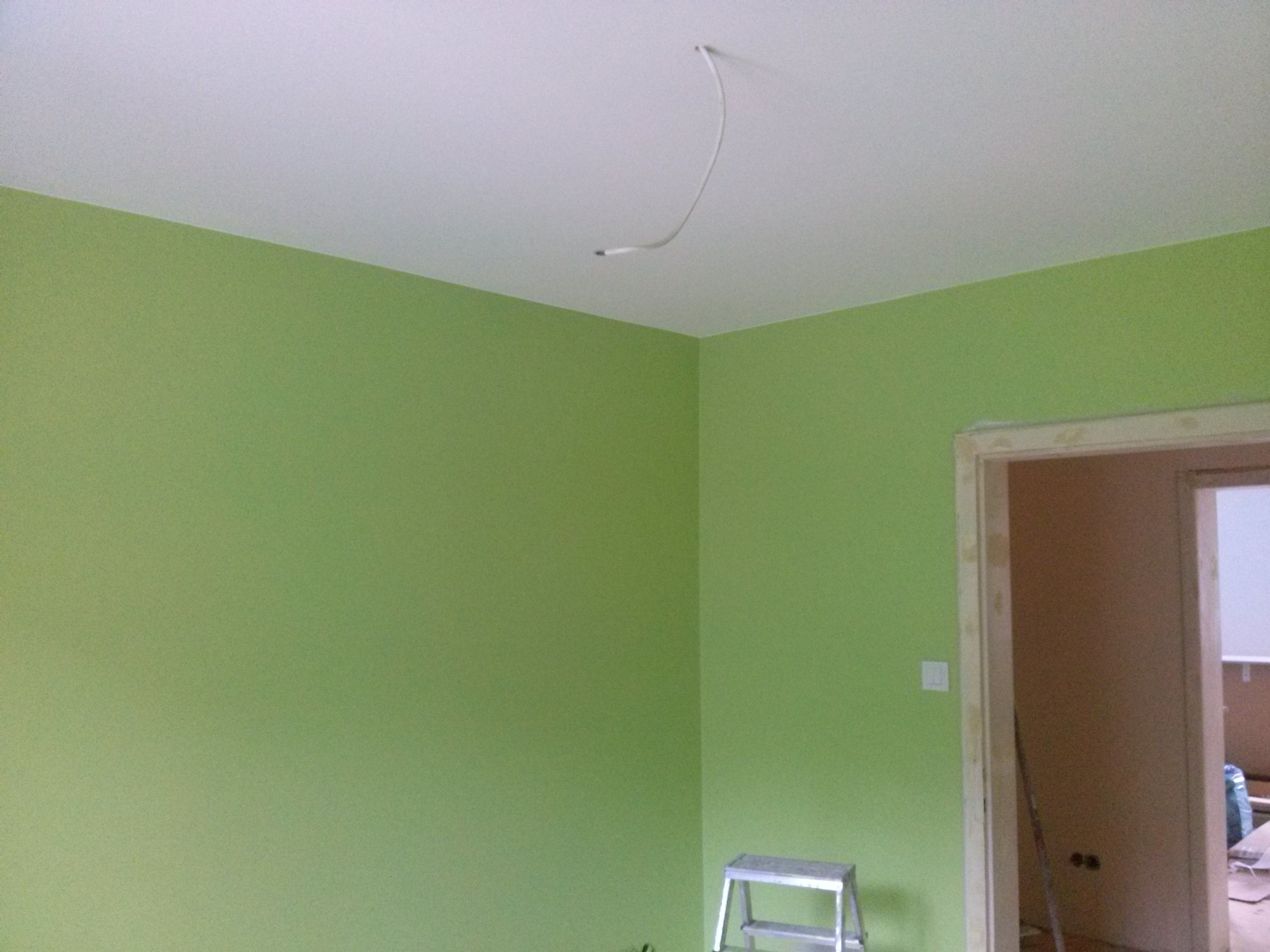 Świeżo pomalowane na zielono wnętrze pokoju z aluminiową drabinką w rogu, widoczne wystające kable z sufitu i otwarte przejście do innego pomieszczenia.